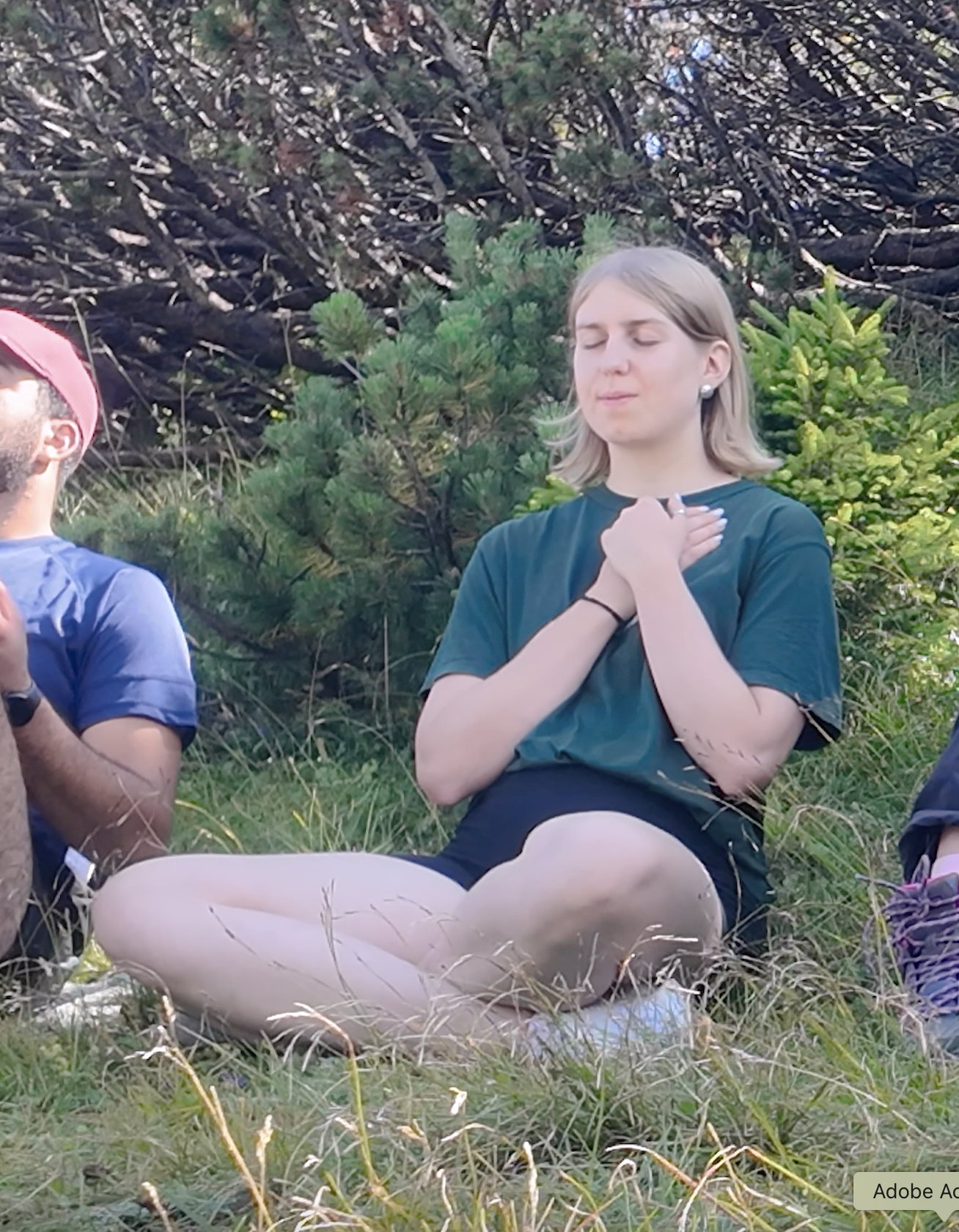 A woman with blonde hair sitting cross-legged outdoors with eyes closed, hands over her heart, surrounded by green trees.