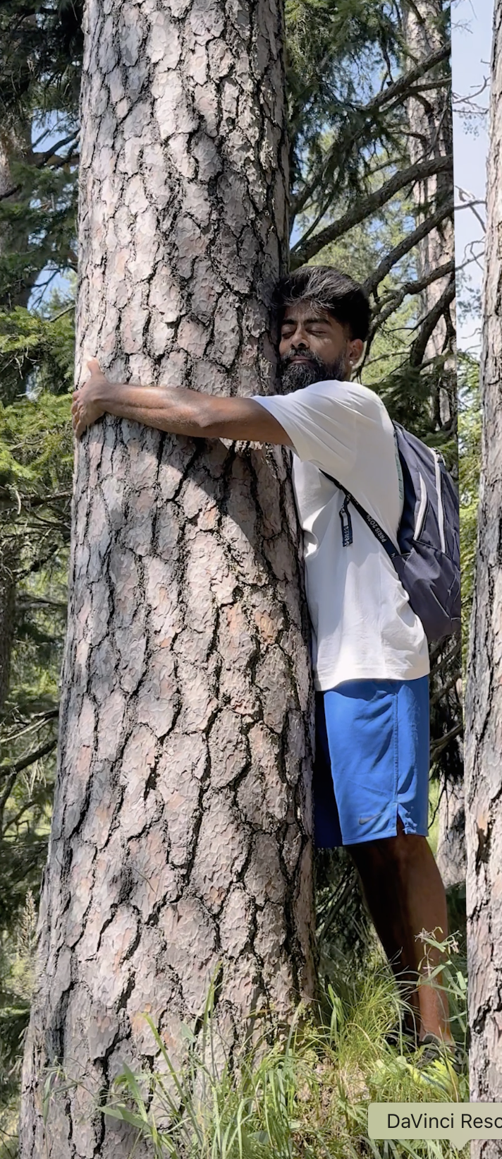 A man with a beard hugging a large tree in a forest, wearing a white t-shirt, blue shorts, and a backpack, with his eyes closed.