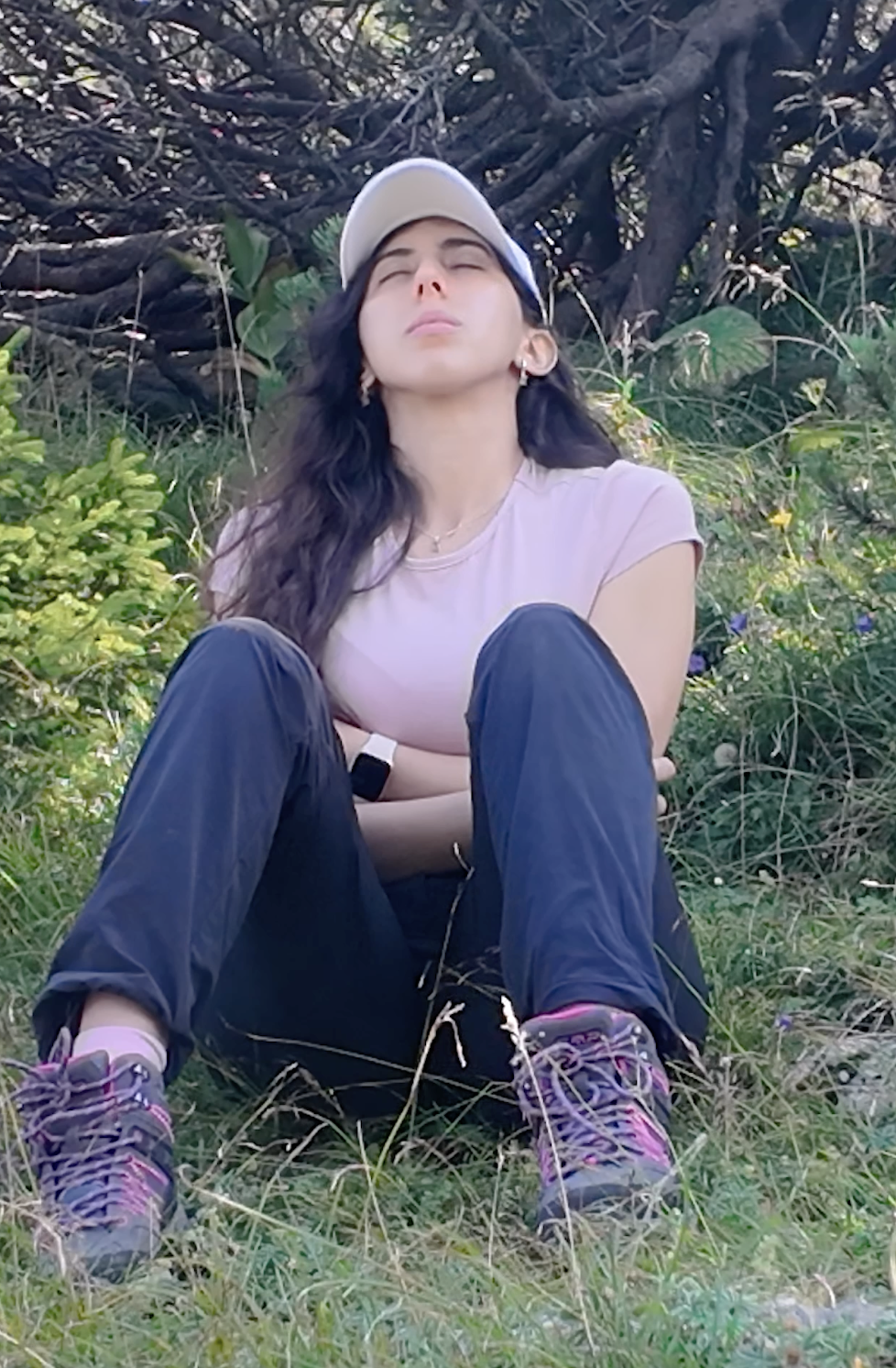 Young woman with long dark hair, wearing a beige cap, pink T-shirt, black pants, and hiking shoes, sitting outdoors in front of a large bush or tree with her eyes closed and arms crossed.