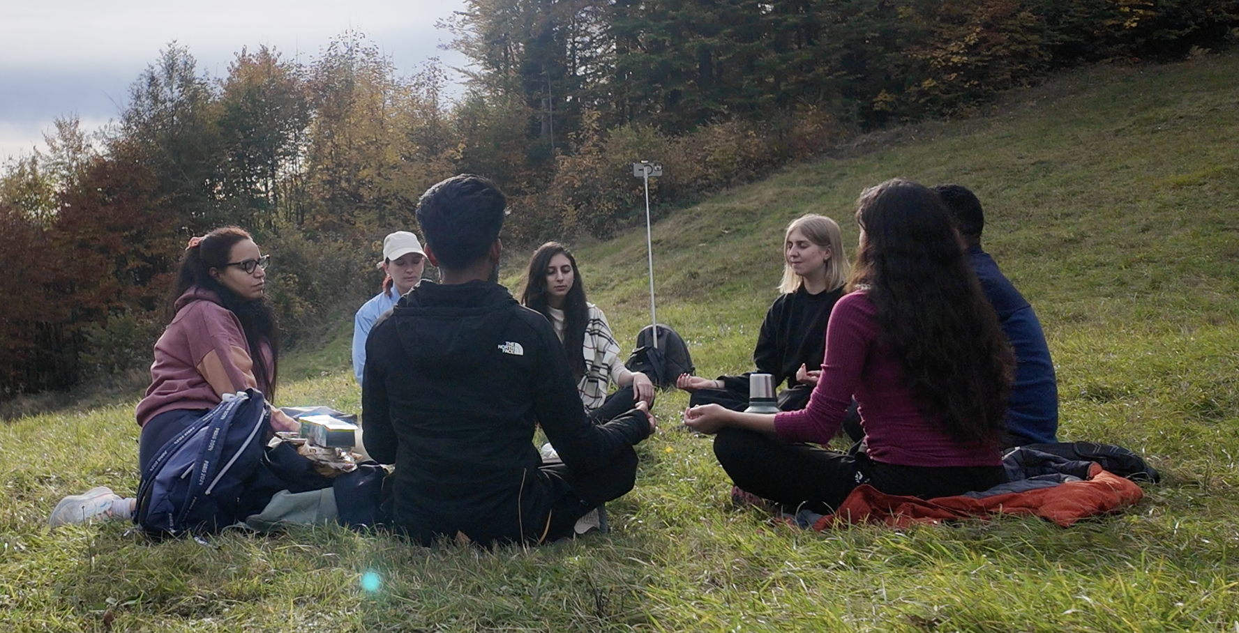 Group of people sitting cross-legged on grass outdoors, participating in an activity with a weather station instrument, surrounded by trees with fall foliage.