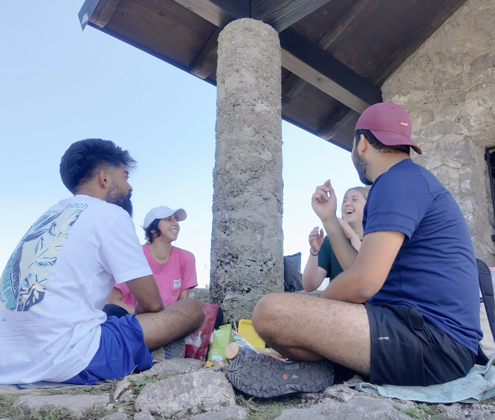 A group of five young people sitting on rocks outside, engaged in conversation and smiling, under a wooden roof supported by a stone pillar, during daytime.