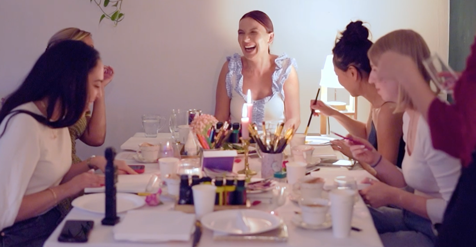 Group of women sitting around a table, laughing and using their phones during a social gathering.