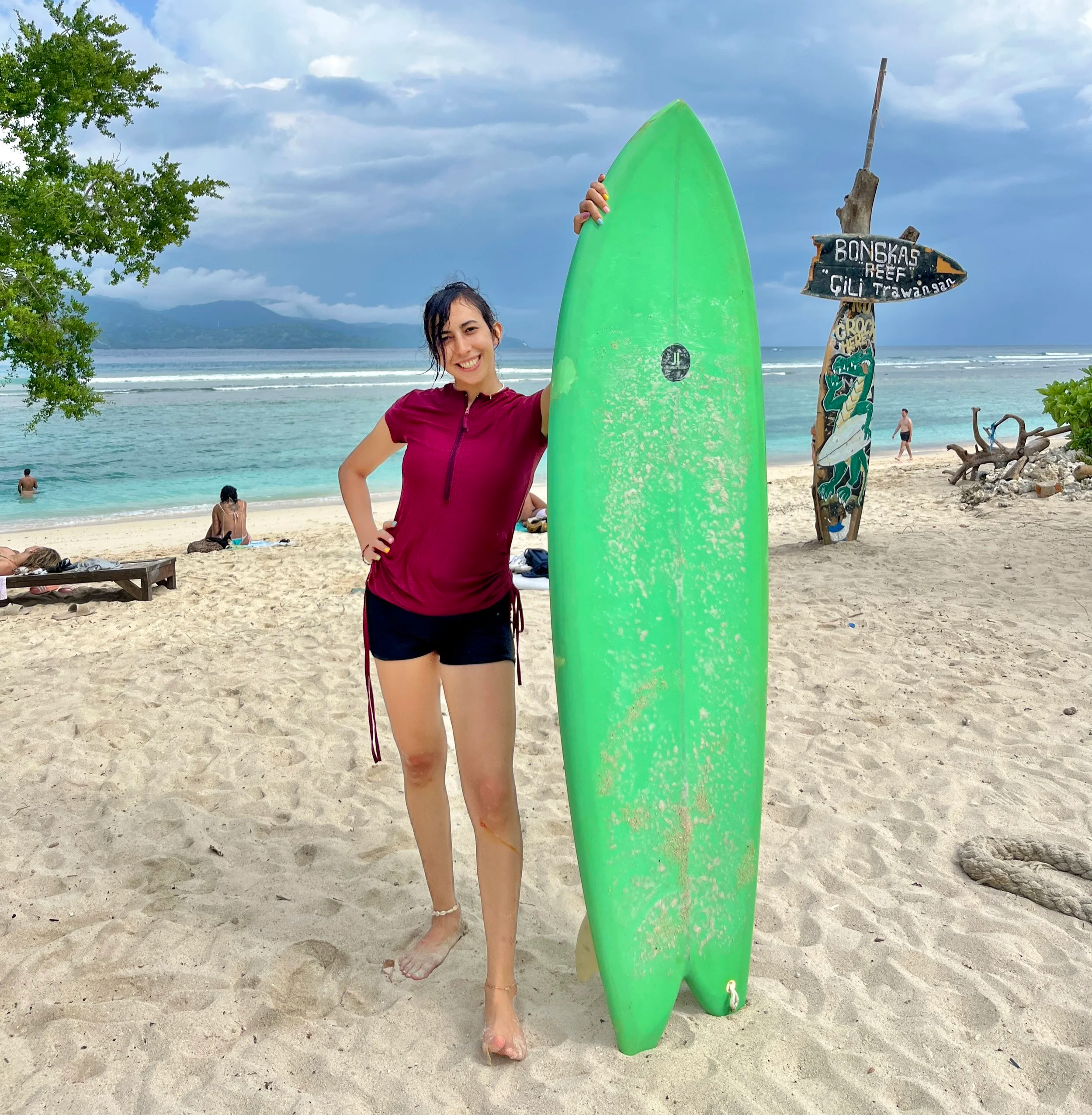 A woman standing on a sandy beach holding a green surfboard, with a cloudy sky and ocean in the background, and a sign pointing towards Bongkas Reef and Gili Trawangan.