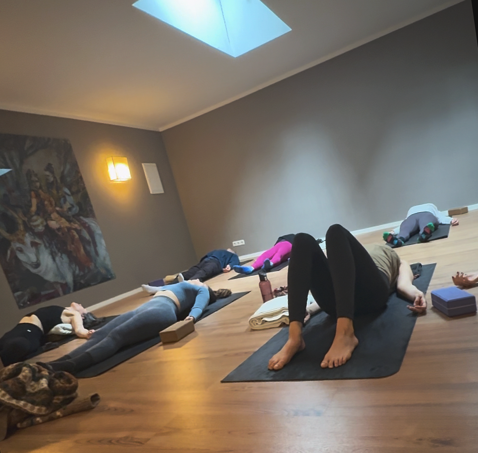 People participating in a yoga class, lying on mats in a dimly lit room with a ceiling skylight and a painting on the wall.