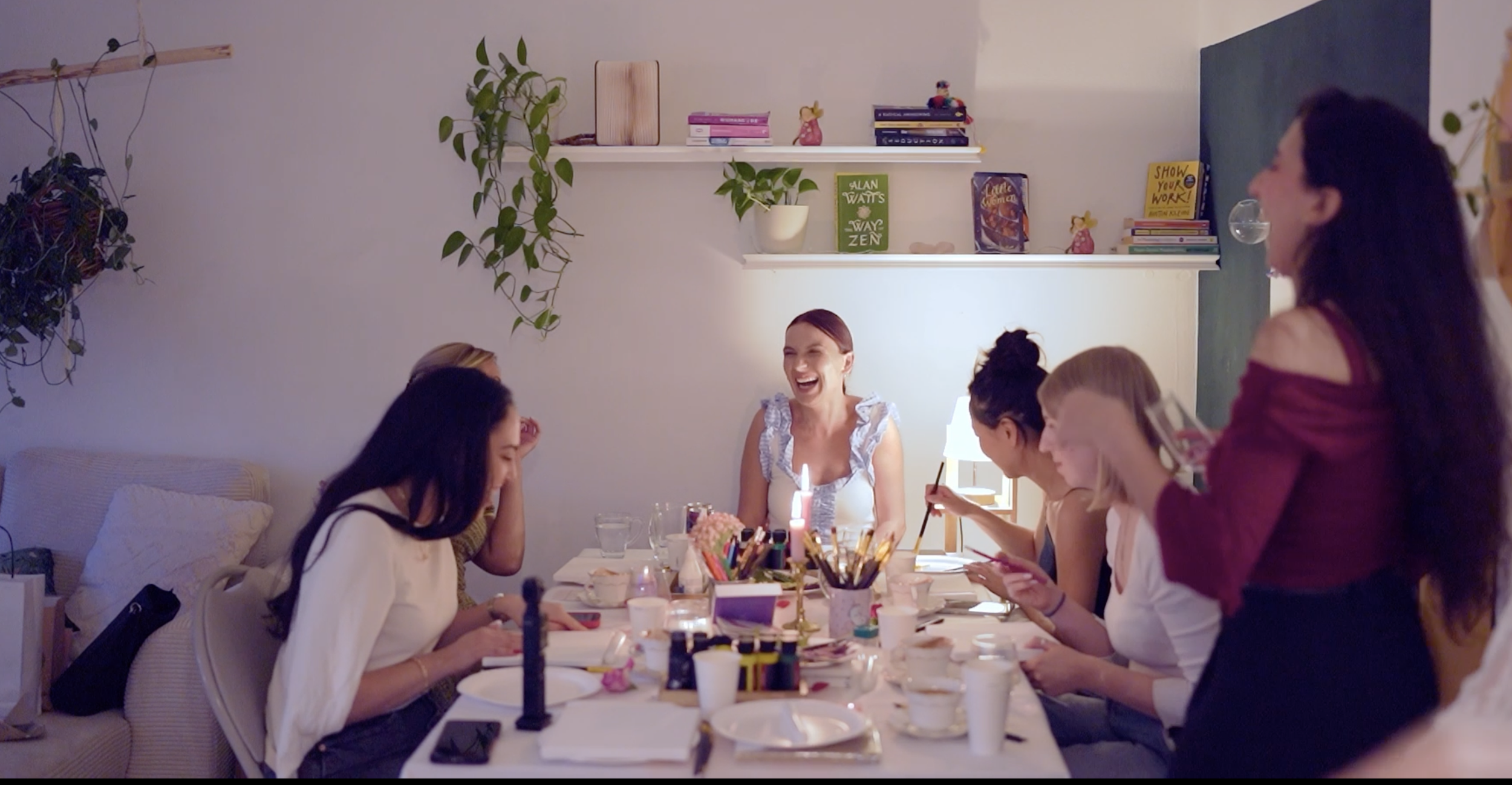 A group of women gathering around a table enjoying a celebration, with some using their phones and one woman smiling and laughing.