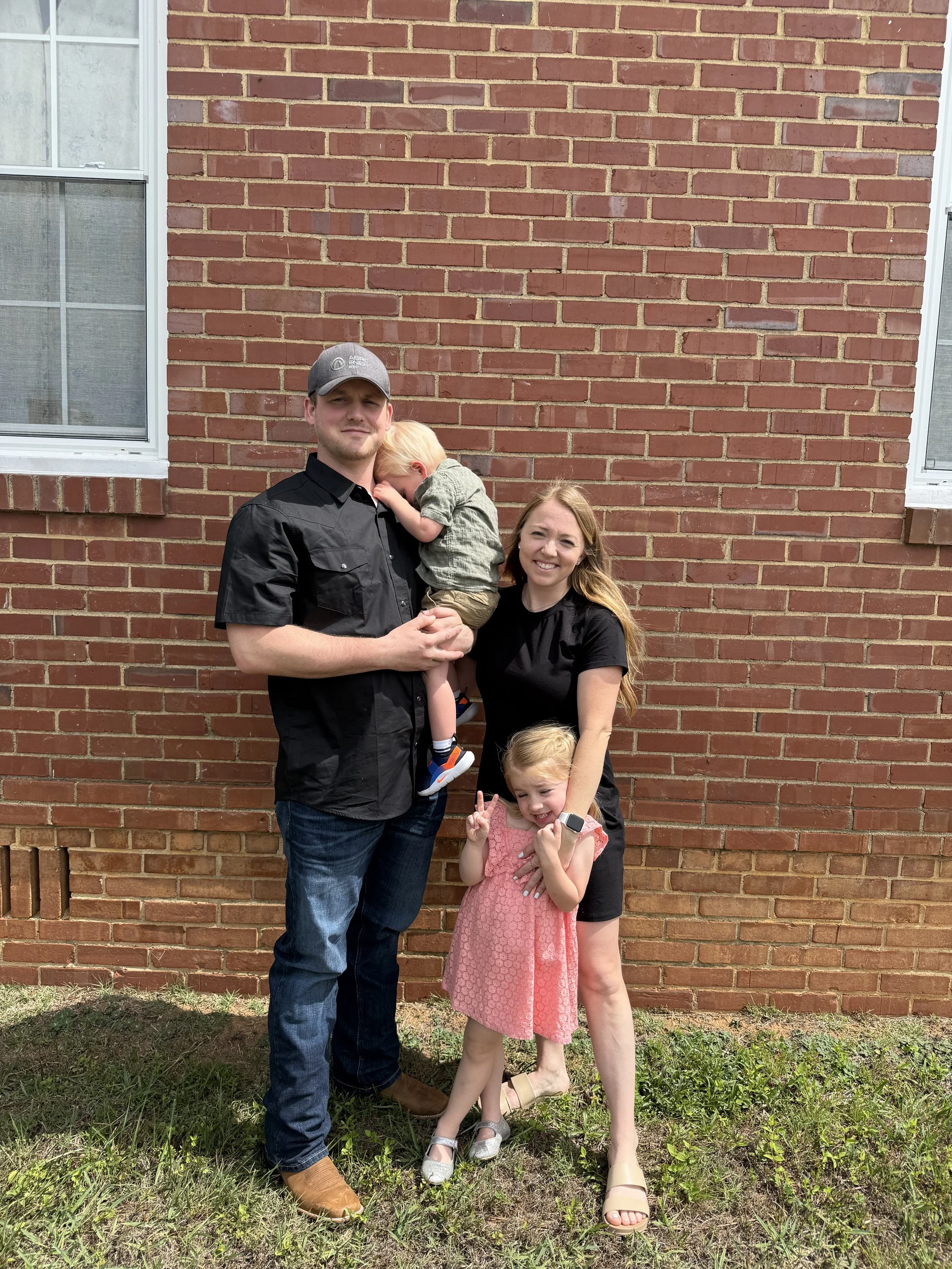 A family of four stands outside in front of a red brick wall. The father, wearing a black shirt, blue jeans, and a gray cap, is holding a young boy with blond hair, dressed in a green shirt and shorts. The mother, with long hair, wears a black shirt and black shorts. She is smiling and standing beside a girl in a pink dress, with long hair, making peace signs with her fingers. The girl is wearing a smartwatch on her left wrist and beige sandals. The ground has grass and dirt.