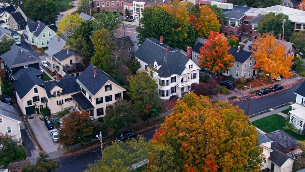 Aerial view of a neighborhood with houses, colorful trees, and a street lined with parked cars during fall.