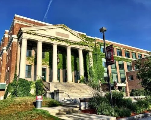 A large brick building with white columns and a decorative pediment, surrounded by green ivy and bushes, with steps leading up to the entrance and a lamppost nearby.