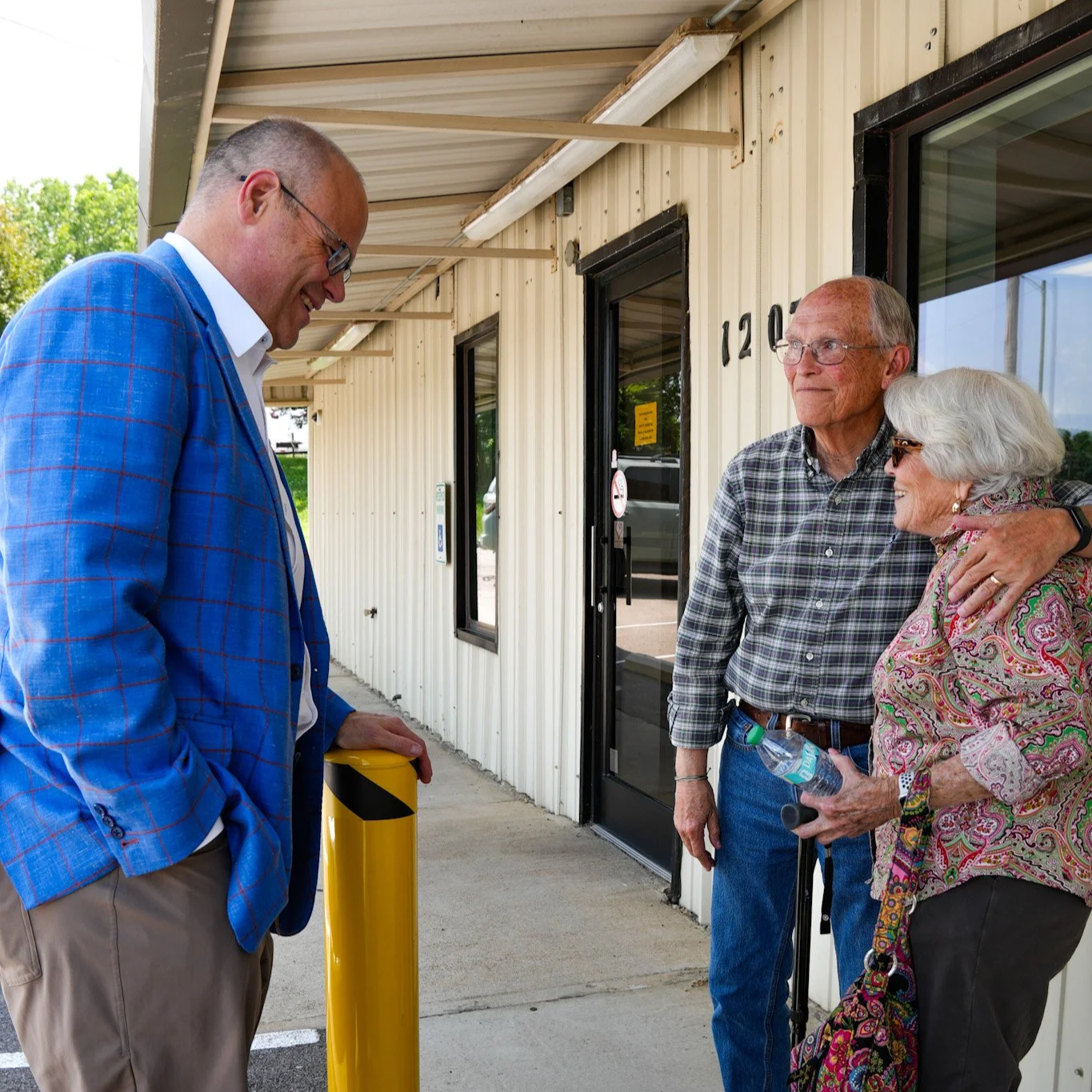 A man in a blue plaid blazer smiles and looks down at a yellow parking bollard, while an elderly couple with a man and woman, the woman wearing sunglasses, embrace in front of a building with the number 120 on it.