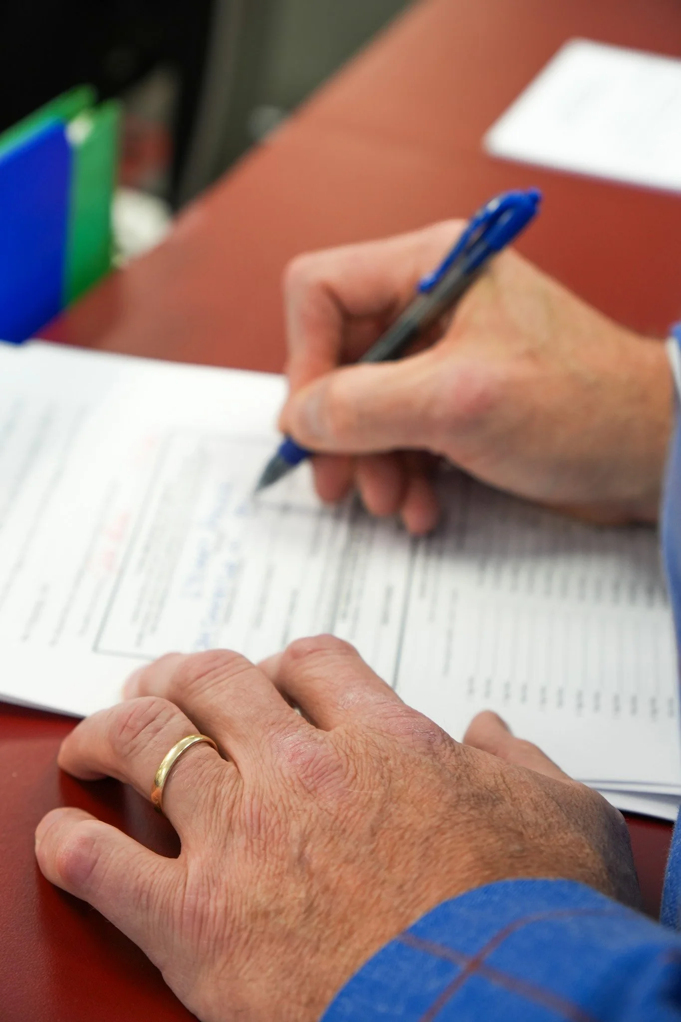 Close-up of a person's hand with a wedding ring, writing on a document with a blue pen. The document is on a red surface, with a blurred background of colorful folders and a white paper.