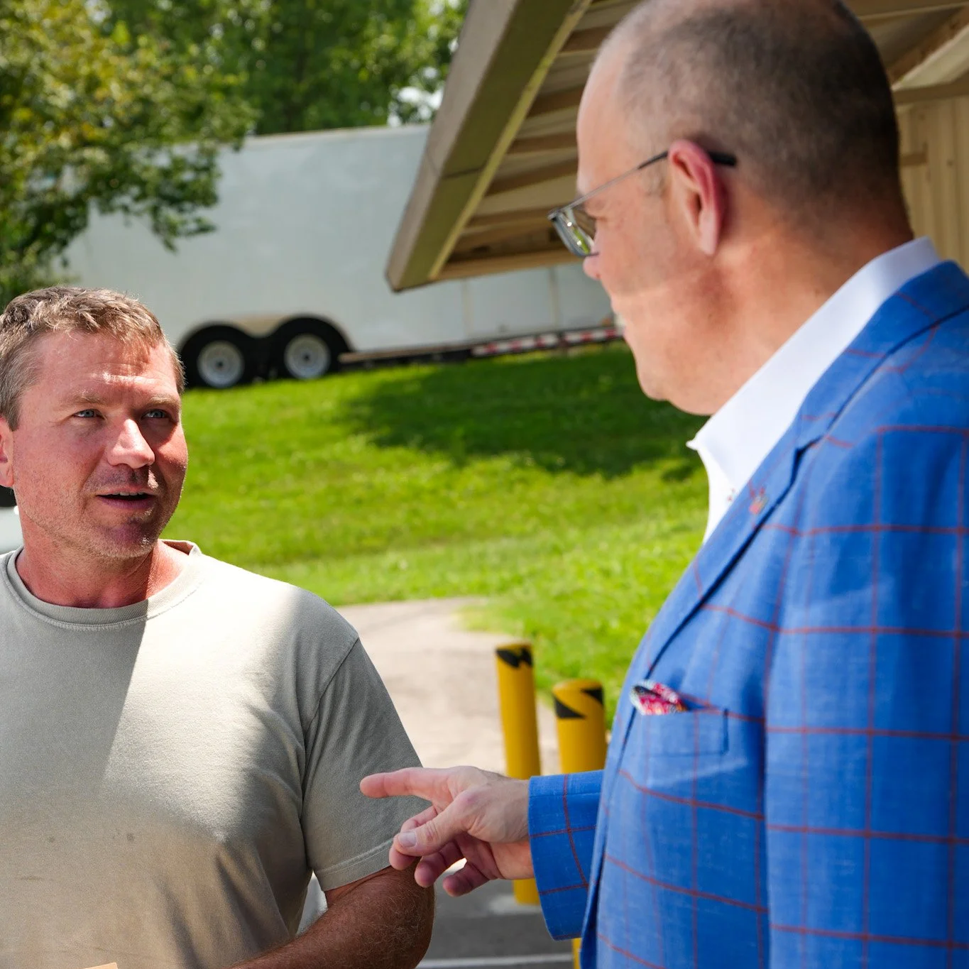 Two men having a conversation outdoors, one in a gray t-shirt and the other in a blue plaid suit jacket and glasses, with green grass and trees in the background.