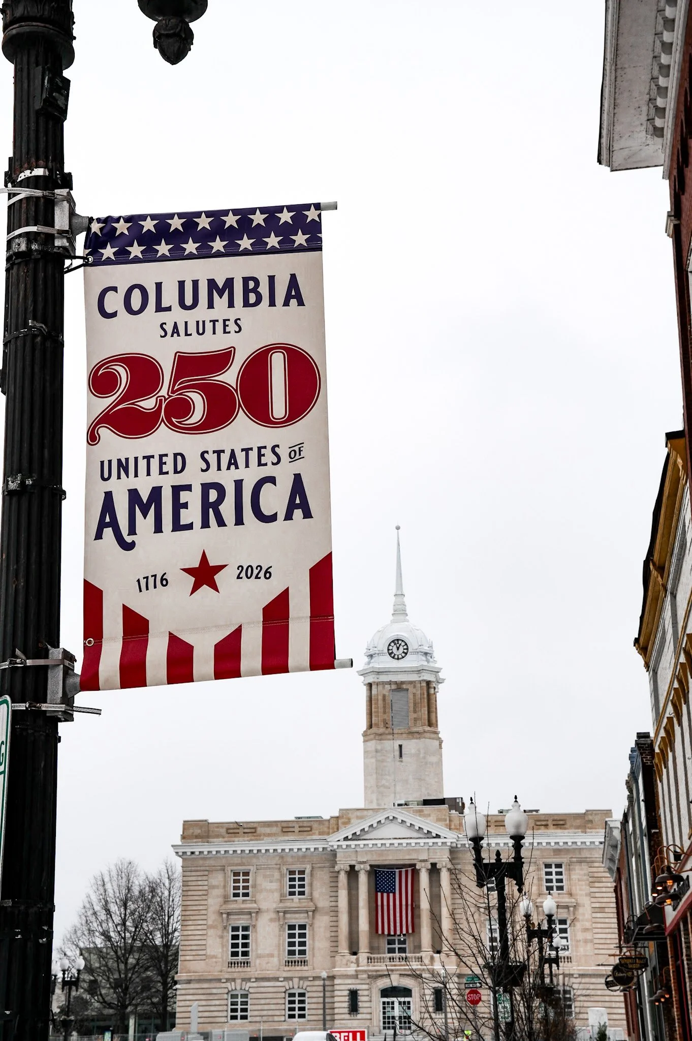 A street scene with a historic government building featuring a clock tower and American flags. A banner celebrating the United States' 250th anniversary is hanging on a lamppost, which also has a flag depicting stars and stripes.