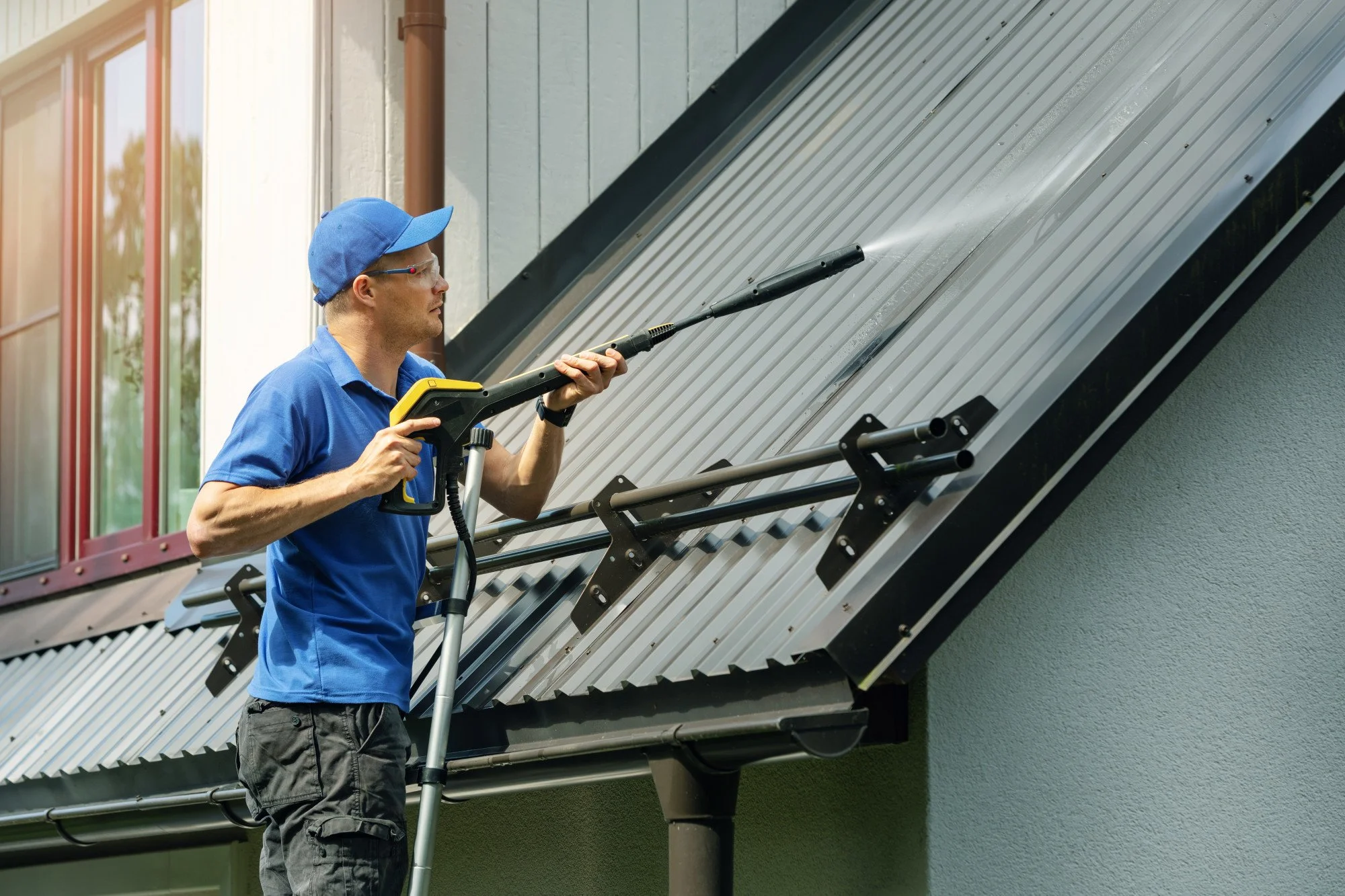 A man wearing a blue shirt, blue cap, and safety glasses is using a pressure washer to clean the metal roof of a house. He is standing on a ladder and focusing on the roof surface.