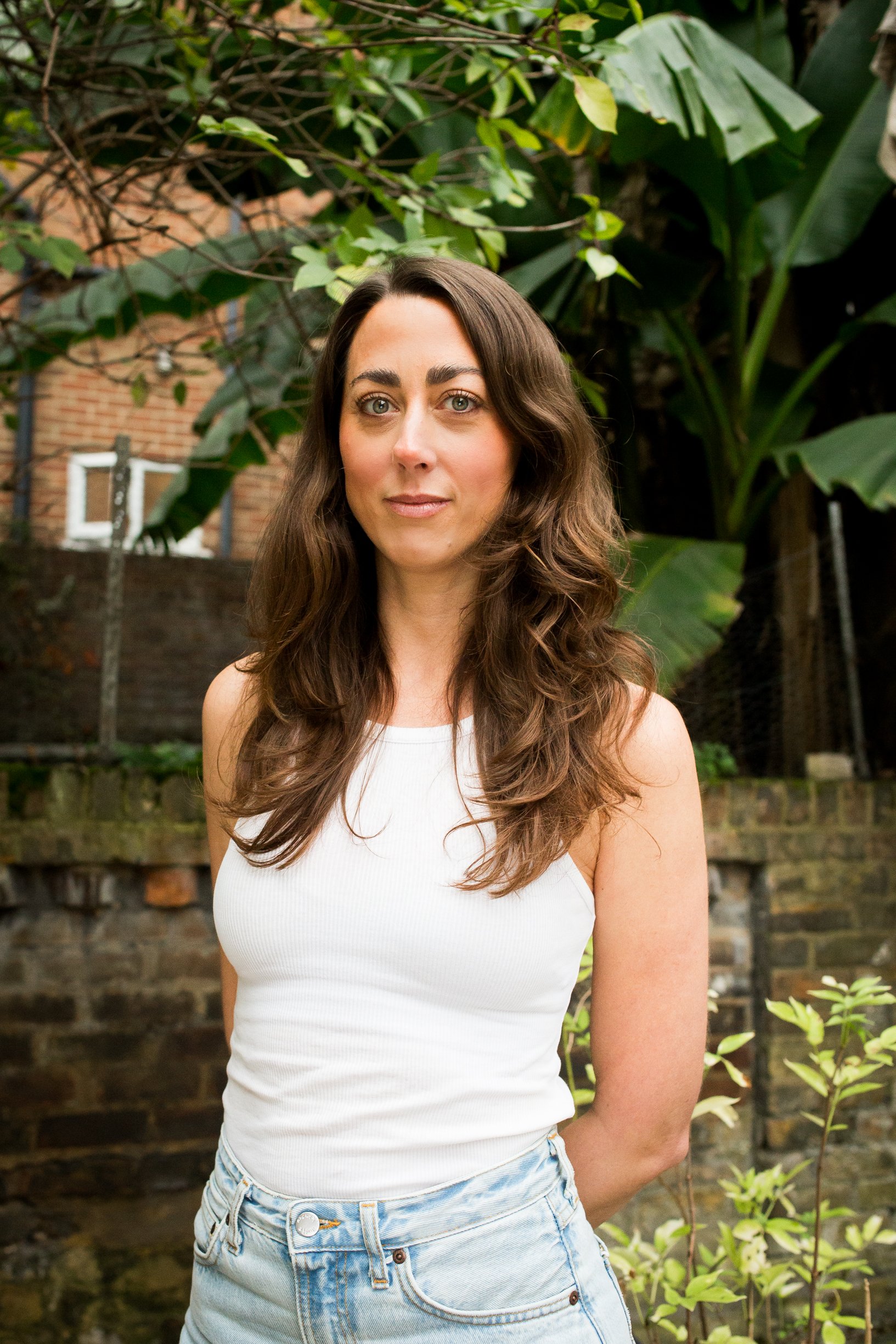 Woman with long brown hair wearing a white top looks at the camera with a friendly expression, standing in front of trees