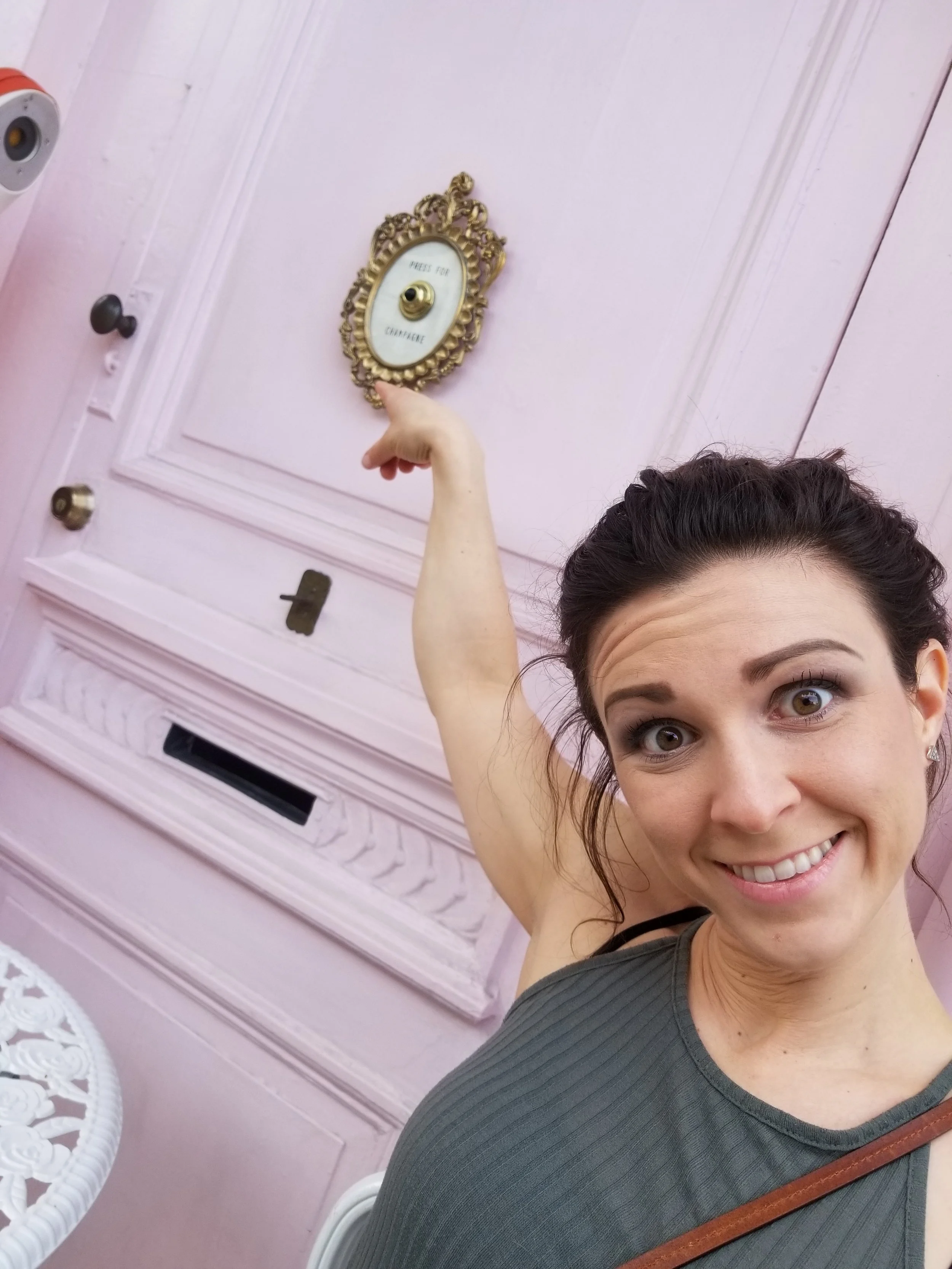 A woman with dark hair in a ponytail smiling at the camera, wearing a gray top, standing outside in front of a pink door with decorative molding and a mirror in an ornate gold frame that indicates to press for champagne.
