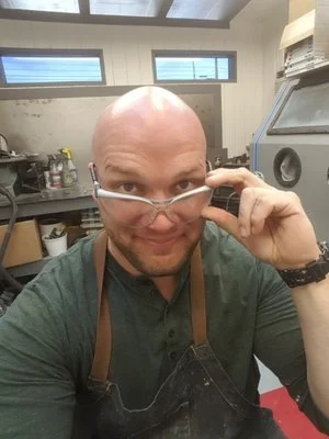 A man with a shaved head and glasses, wearing a green shirt and apron, smiling in a workshop or laboratory setting.