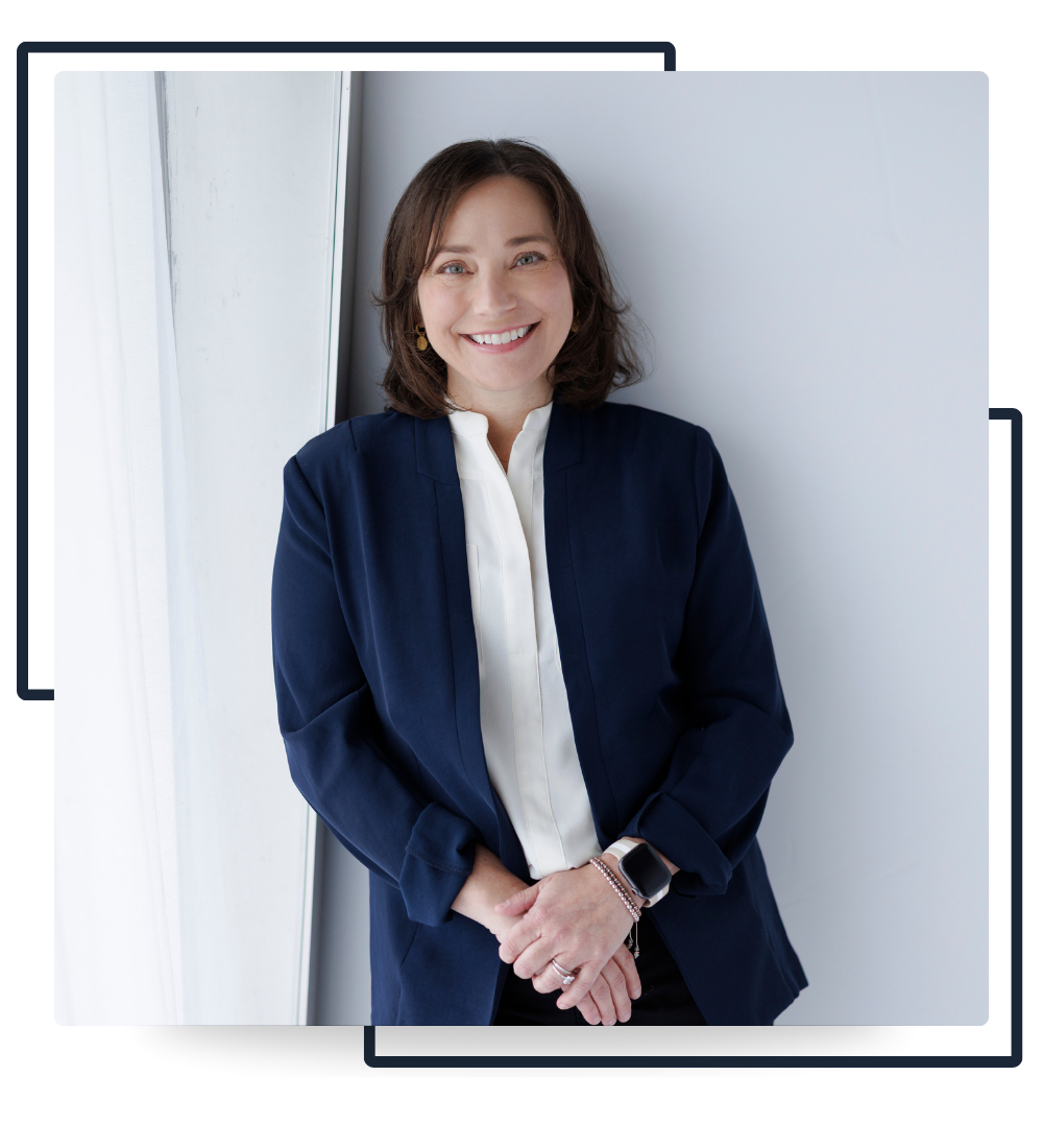 A smiling woman with brown hair, wearing a navy blue blazer, white blouse, and a smartwatch, standing by a window.