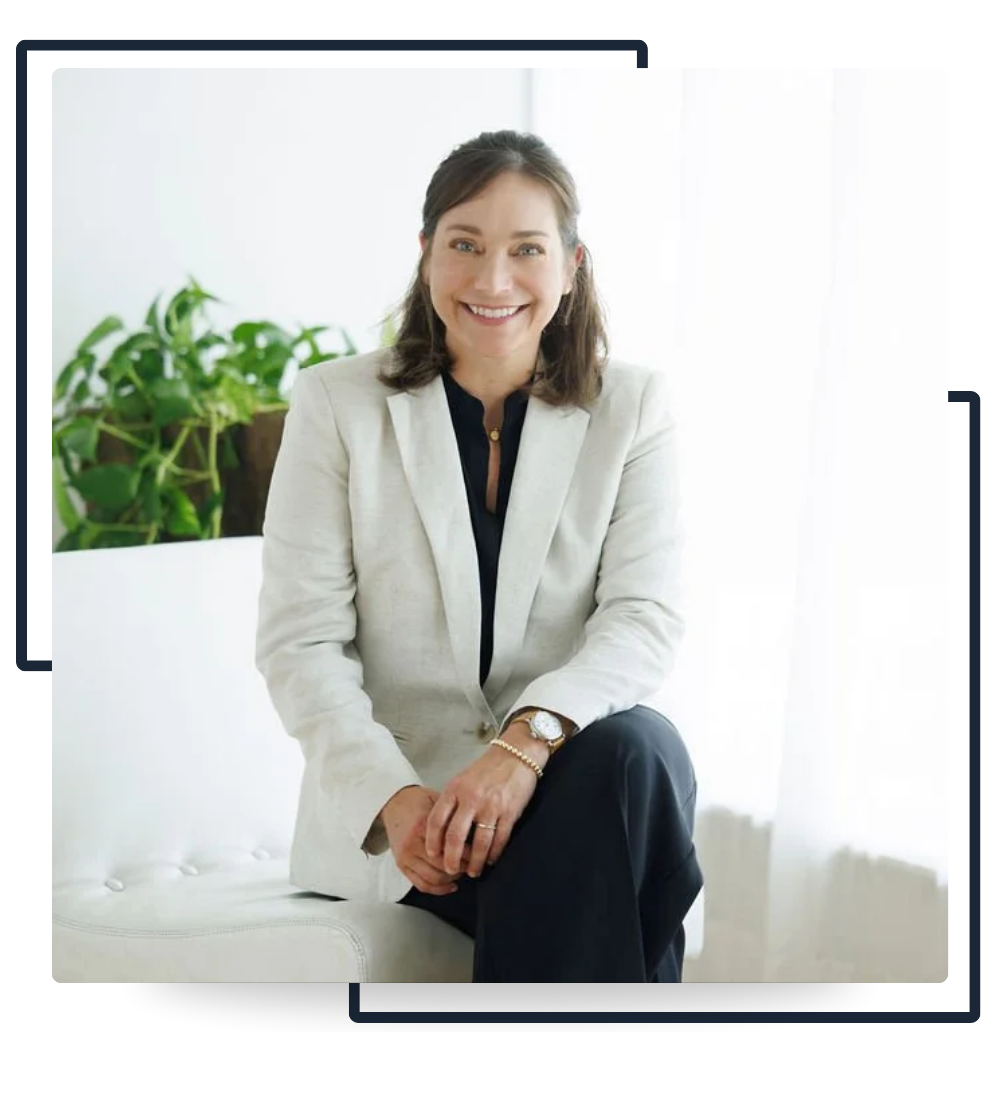 Farrah Chrstos, LCSW, LISW in a white blazer and black pants sitting on a white chair, smiling. There is a green plant behind her on a wooden table.