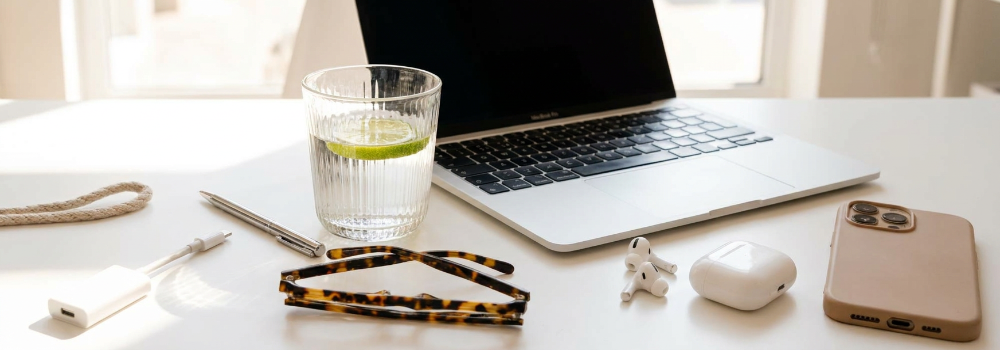 Bright image of a laptop, glass of water with lime, and tortoise glasses. A pair of earbuds is also on the desk.