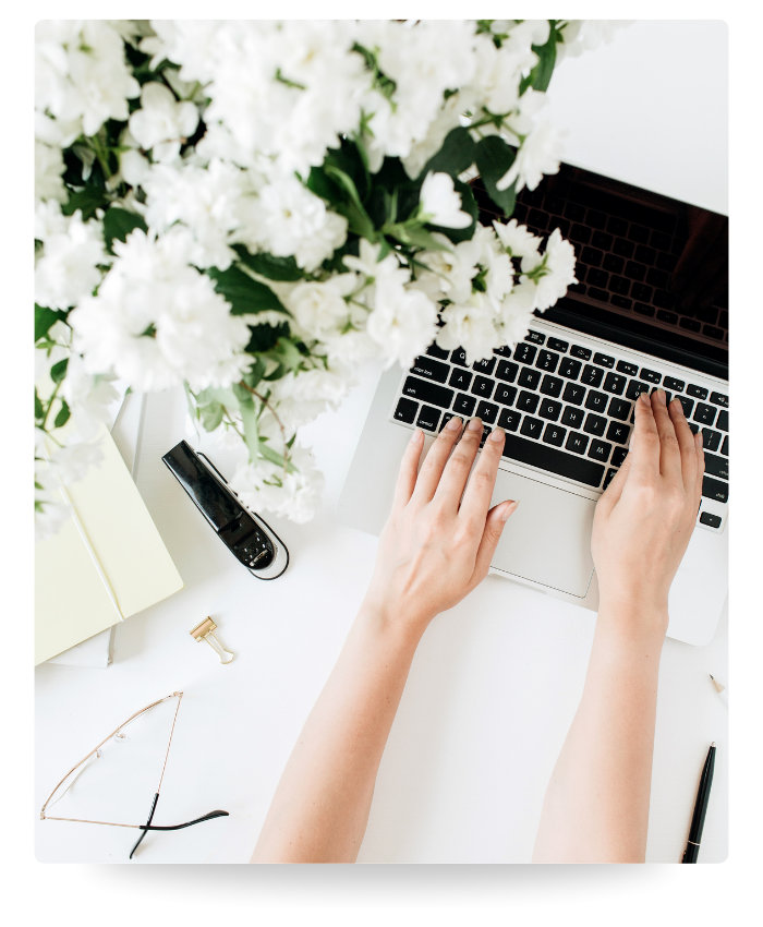 Person typing on a laptop keyboard with white flowers and office supplies on a white desk.