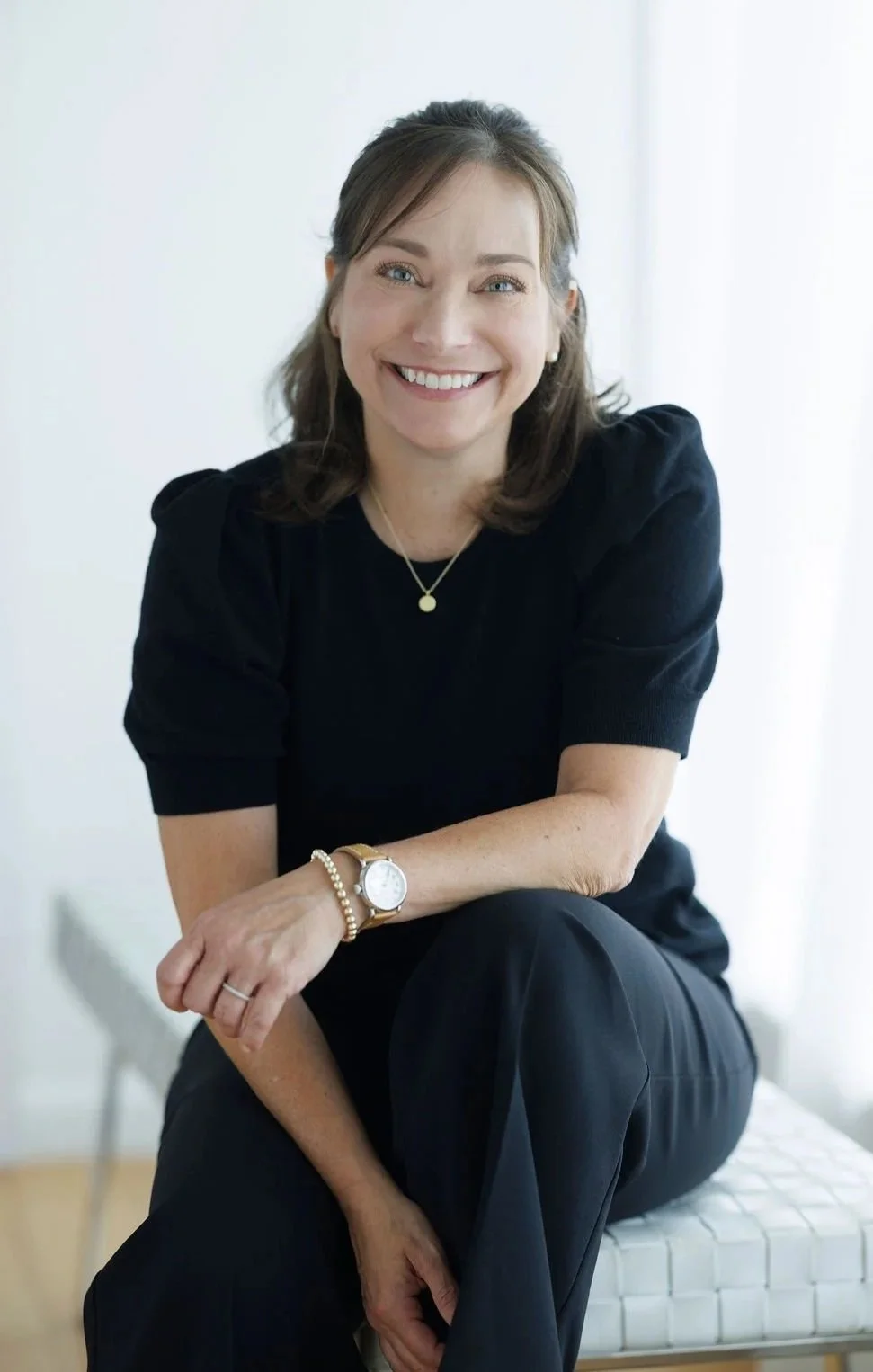 Farrah Chrstos, LCSW, LISW shoulder-length brown hair, wearing a black short-sleeved top, black pants, a watch, a bracelet, and a necklace, smiling and sitting on a white bench in a bright room.