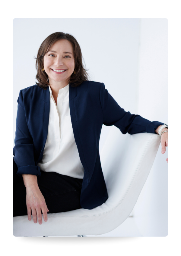 A woman with shoulder-length brown hair, wearing a white blouse and a navy blazer, smiling and sitting on a white modern chair in a bright, minimalistic room.