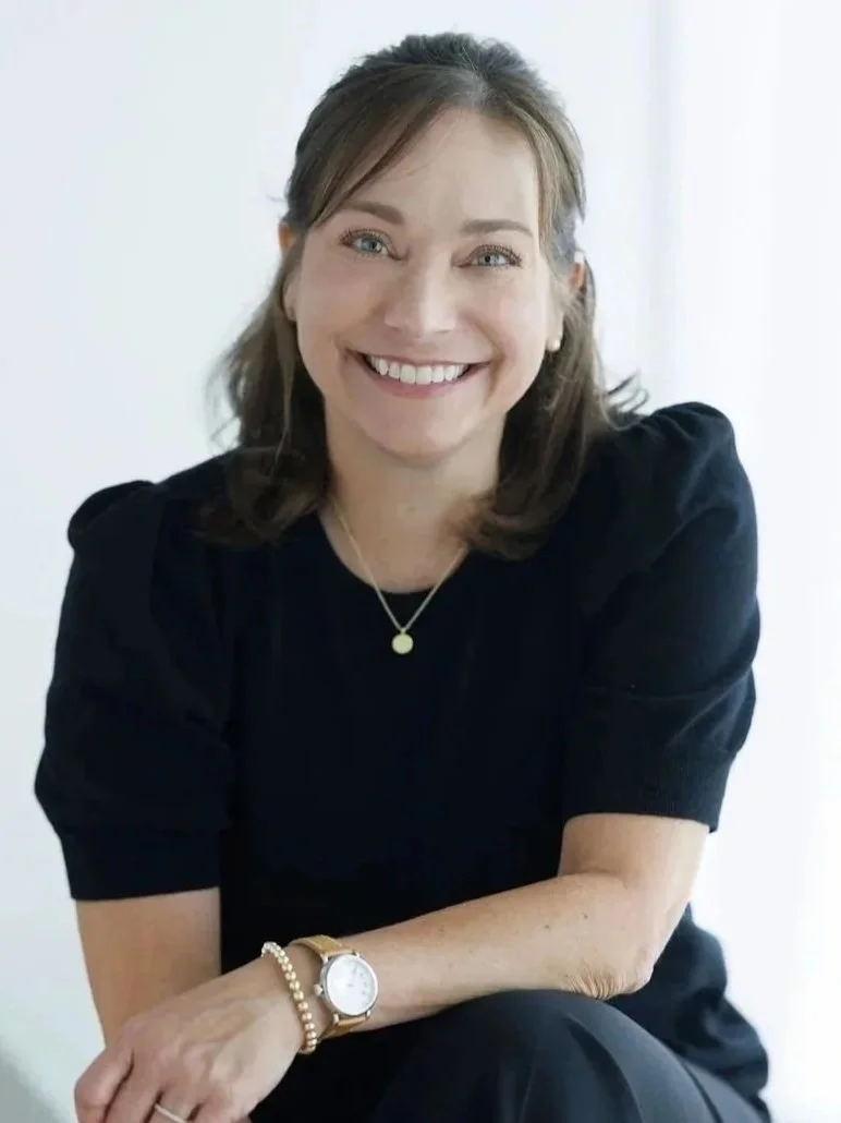 Farrah Chrstos, LCSW, LISW with shoulder-length brown hair and fair skin smiling at the camera, wearing a black top, a wristwatch, and jewelry, sitting against a plain white background.