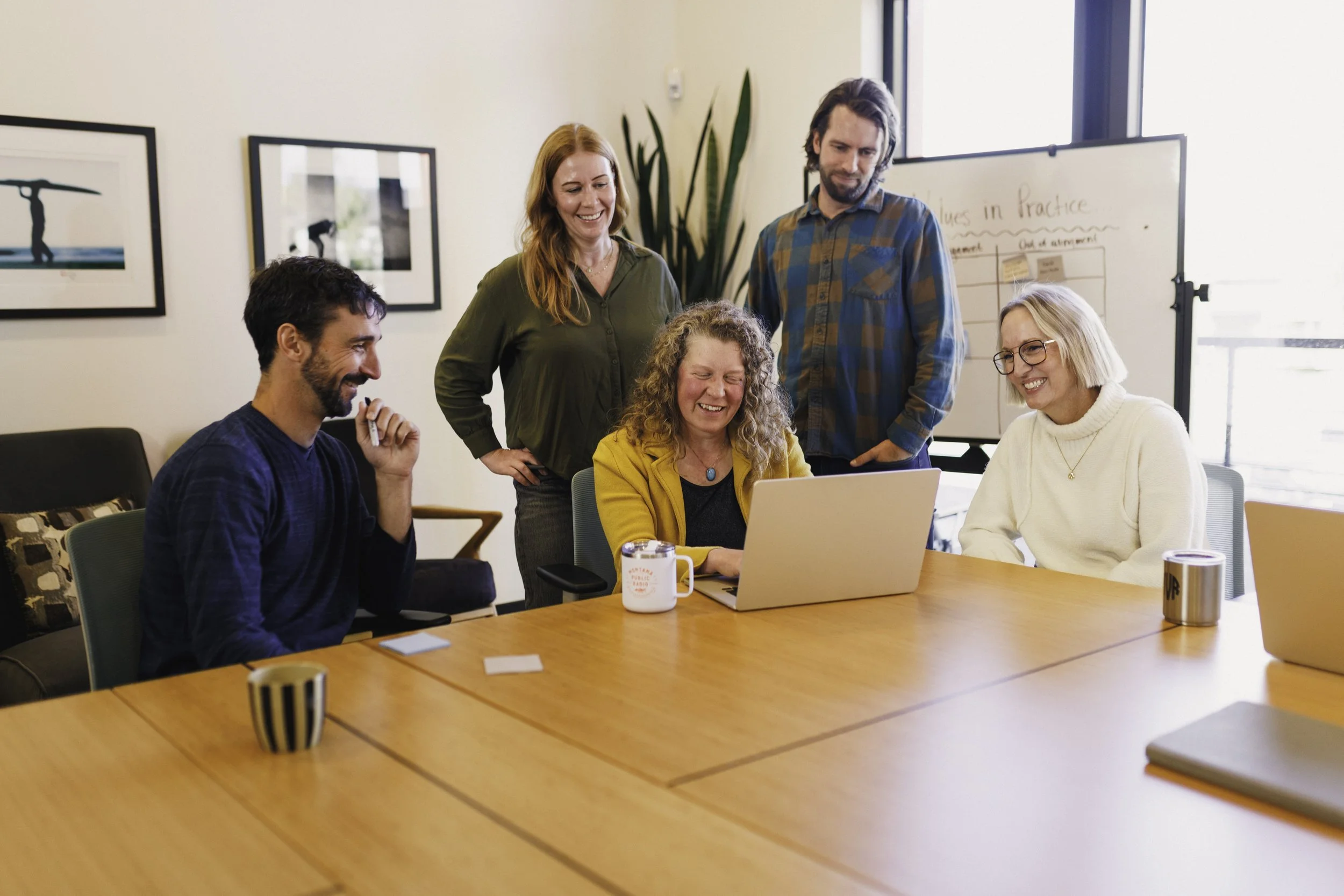 Five coworkers gathered around a laptop at a conference table, smiling and engaging in a meeting in a well-lit office with framed art on the walls.