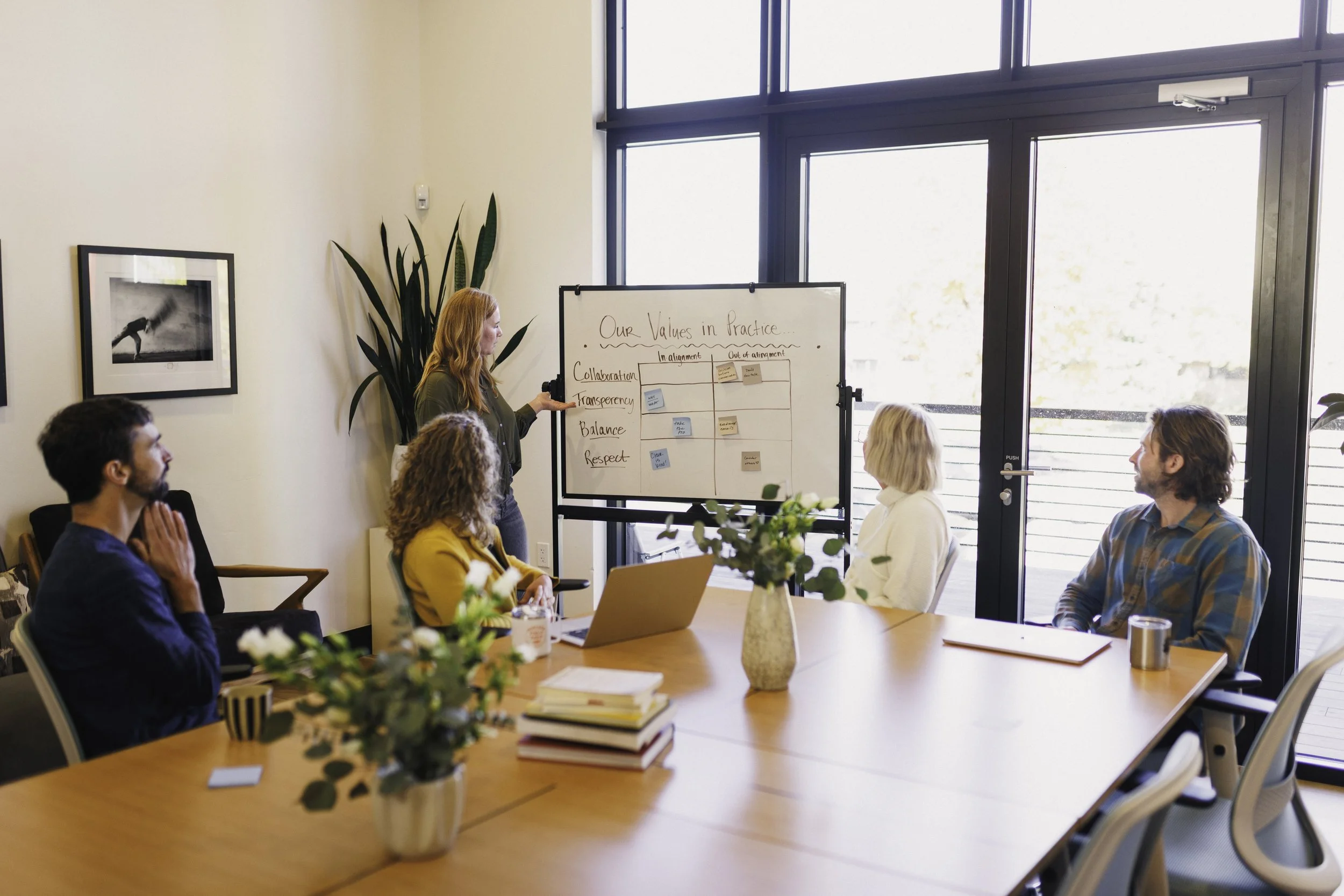 A group of people gathered around a conference table listening to a woman giving a presentation. The presenter stands next to a whiteboard with writing on it, and large windows let in natural light. The table has notebooks, a laptop, and a flower arrangement.