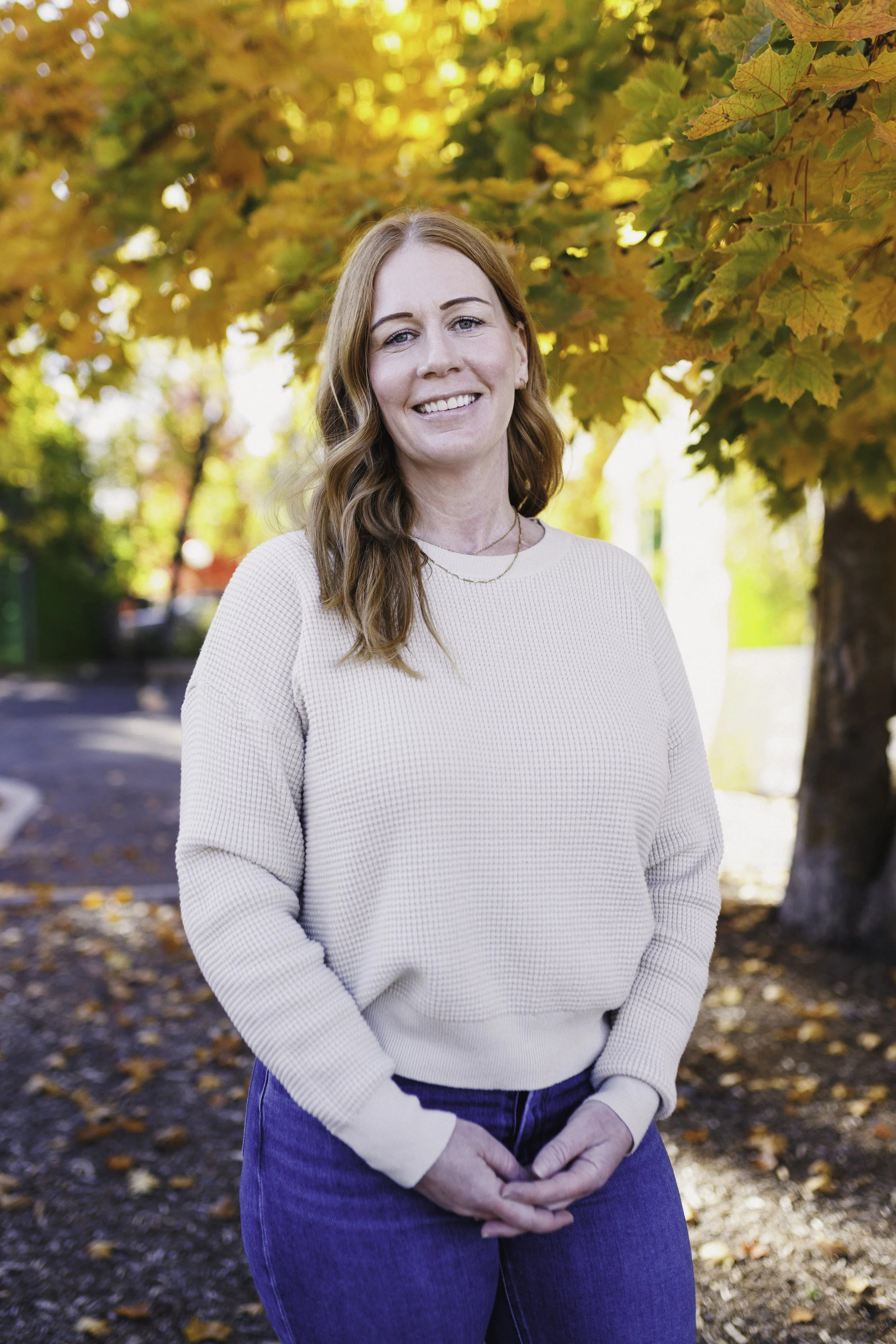 A woman with red hair smiling outdoors in front of autumn leaves during daytime.