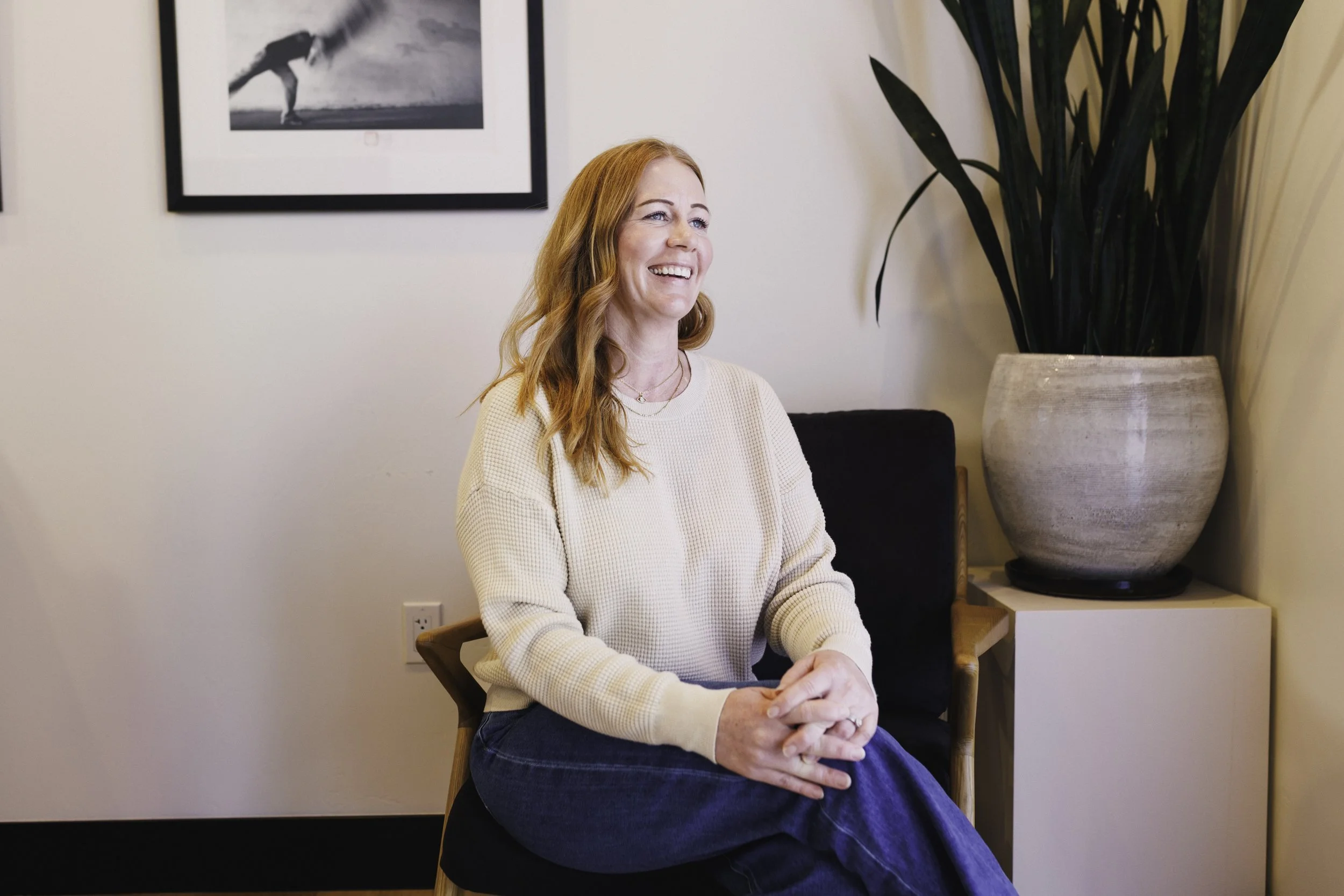 A smiling woman with red hair, wearing a cream-colored sweater, sitting on a chair in an indoor setting with framed black-and-white artwork and a large potted plant behind her.