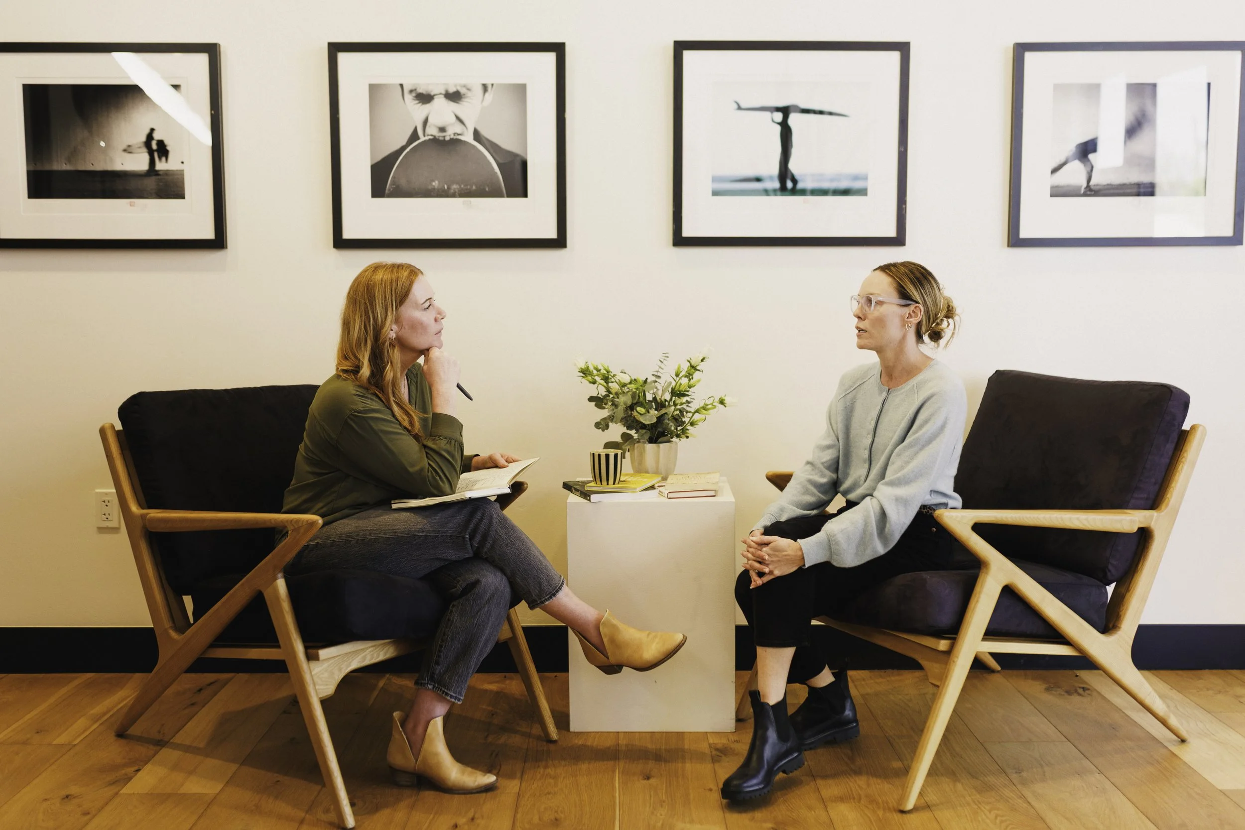 Two women sit and converse in a room with black and white framed photos on the wall. One woman with long red hair and glasses, wearing a light-colored top, listens attentively. The other woman with long red hair, dressed in dark clothing, holds a paper and pen. There is a small table with books and a potted plant between them, and wooden chairs with armrests.