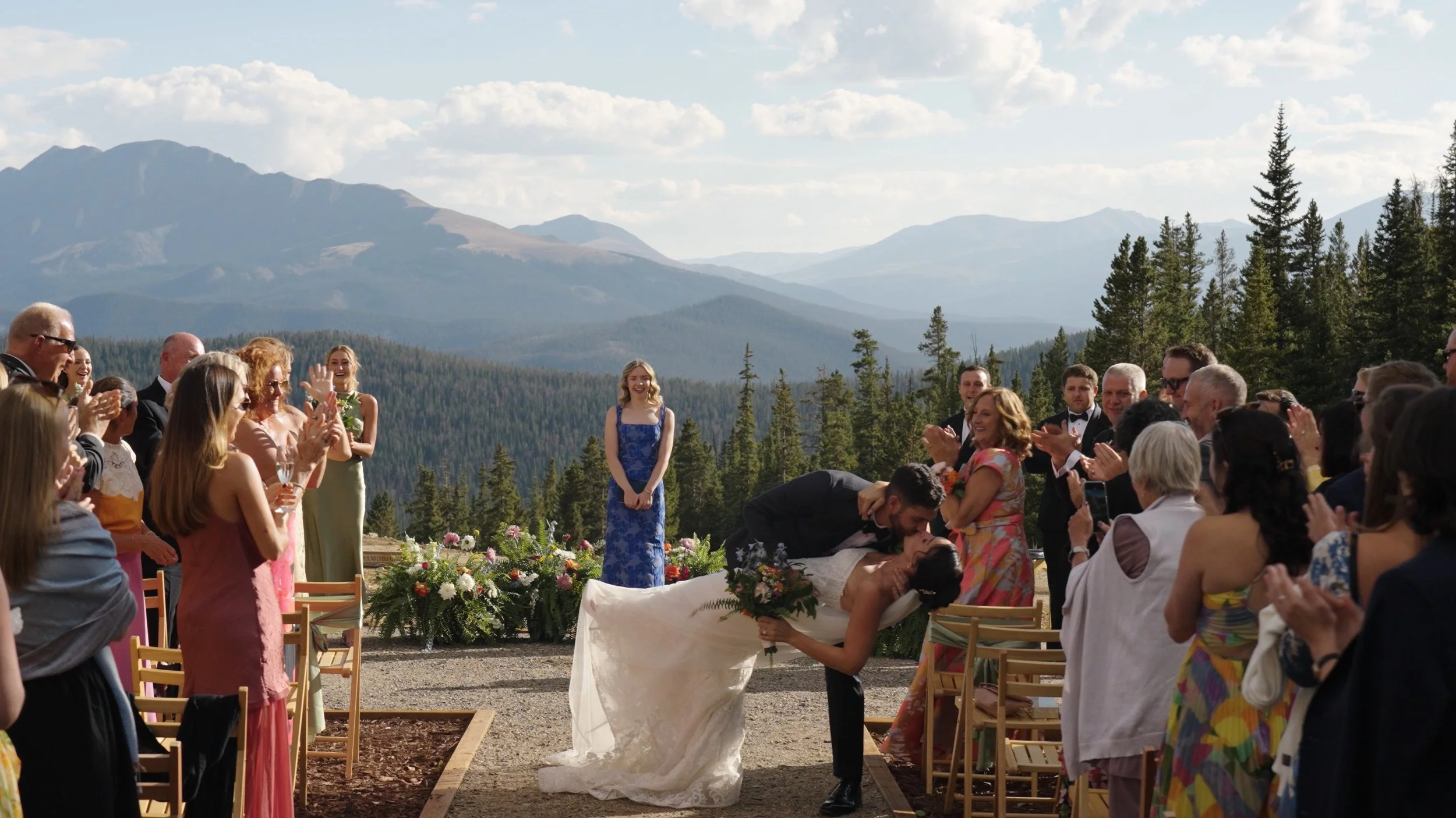 A wedding ceremony taking place outdoors with a mountain and forest backdrop, with the bride and groom kissing while surrounded by guests who are clapping and smiling.