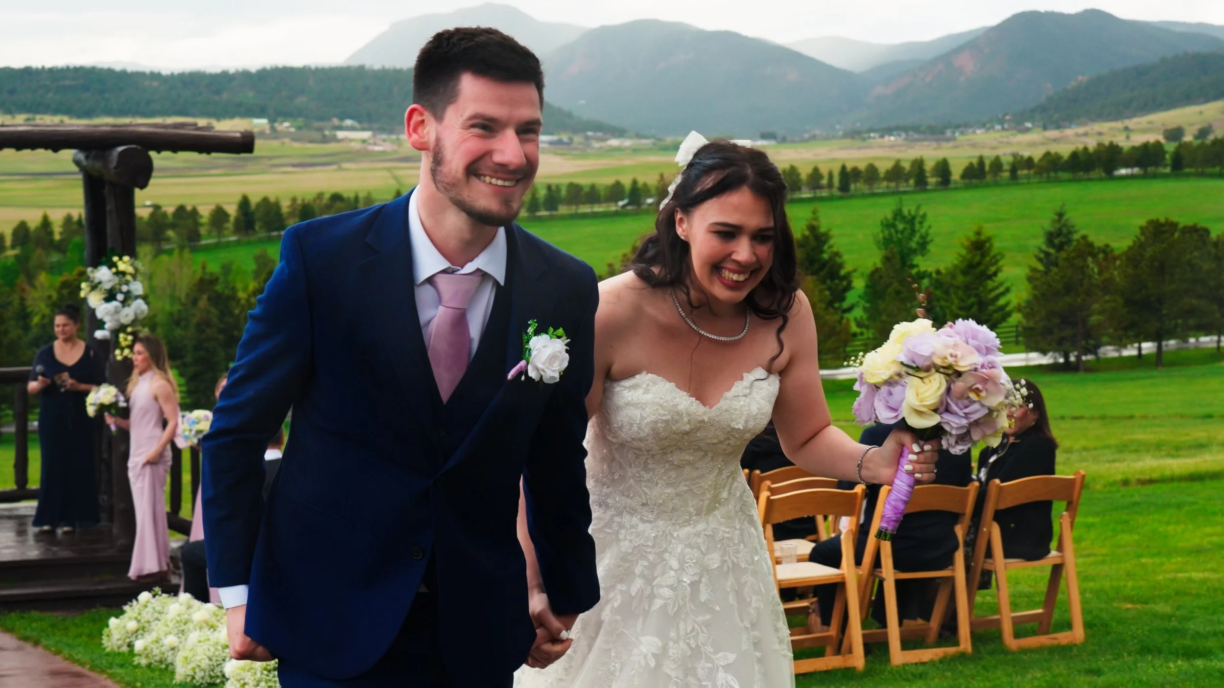 A smiling bride and groom holding hands at an outdoor wedding ceremony with mountains in the background. The bride is wearing a wedding dress and holding a bouquet of pastel-colored flowers, while the groom is dressed in a navy suit and pink tie.