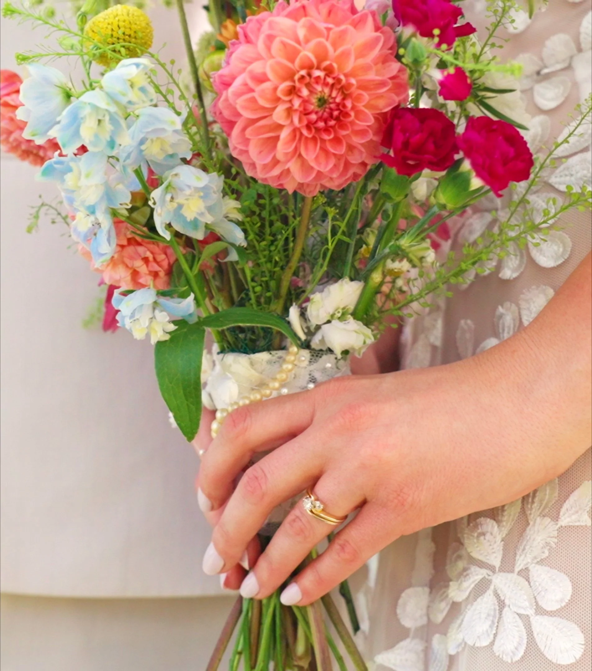 A person holding a colorful bouquet of flowers, including a large pink dahlia, light blue delphiniums, pink and red carnations, and other greenery. The person is wearing a pearl and gold ring and a gold band ring, and a pearl necklace, with a lace fabric visible in the background.