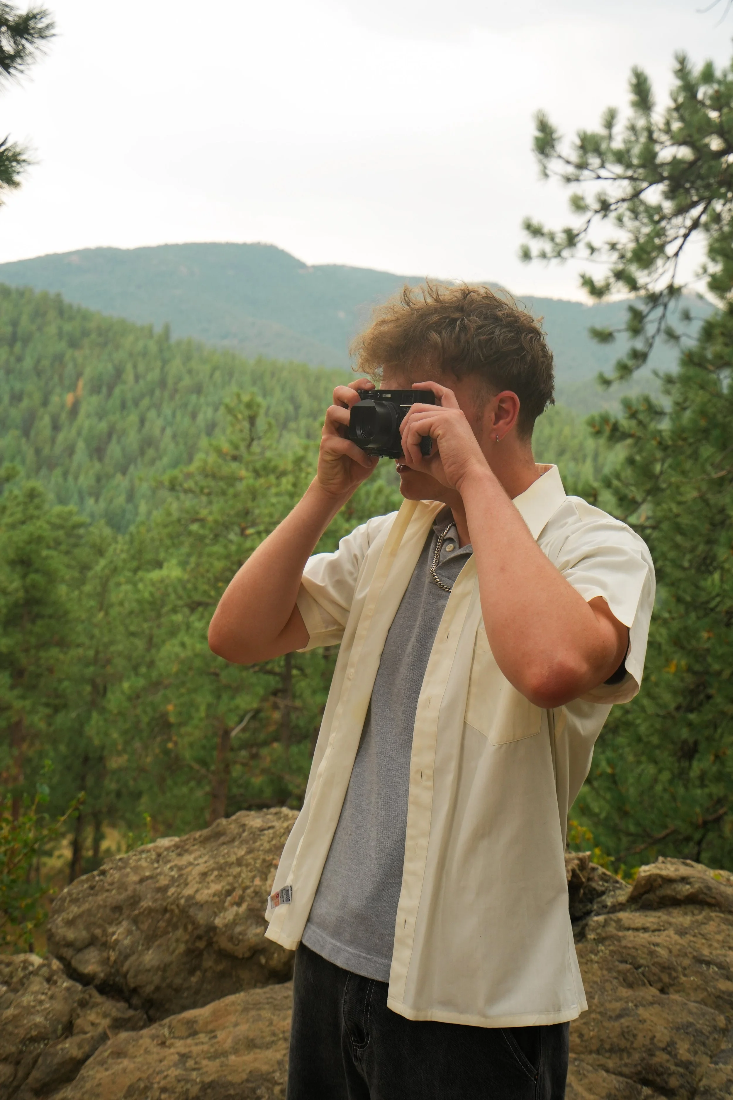 A young person with curly hair takes a photo with a camera outdoors in a mountainous, forested area.