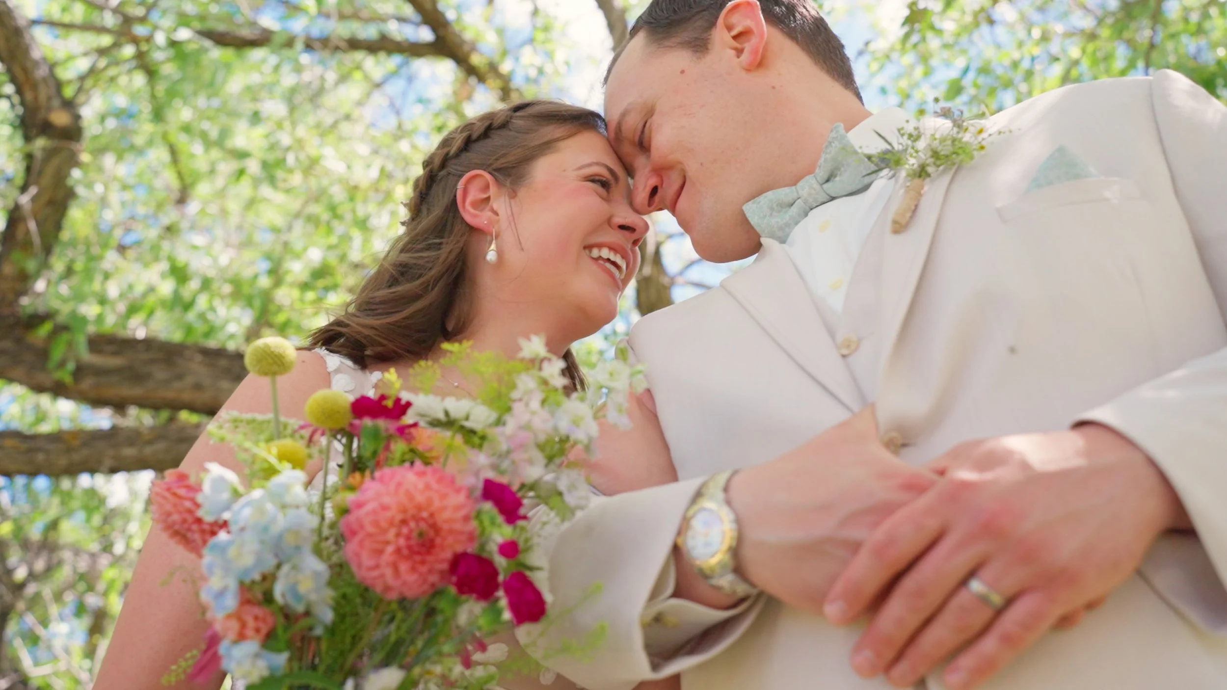 A bride and groom share a close, intimate moment outdoors, touching foreheads while smiling, with the bride holding a colorful bouquet and surrounded by green trees.