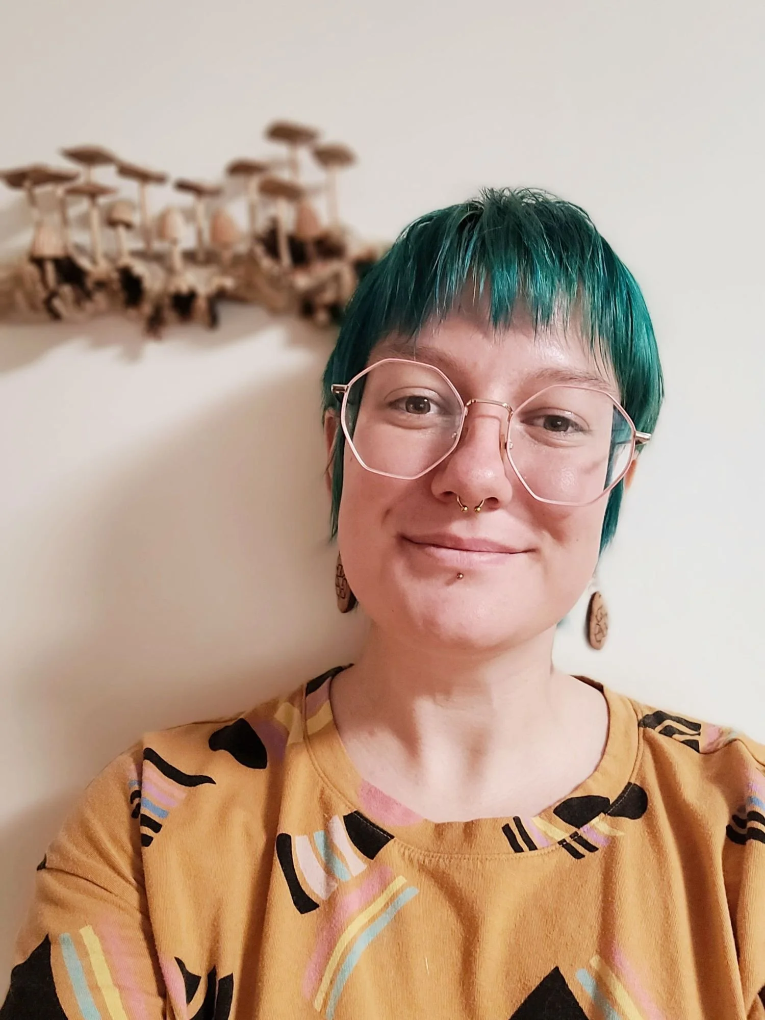 A person with short, teal hair and gold hexagonal glasses smiling. They have a nose piercing and are wearing a mustard-colored shirt with abstract colorful patterns. A wall with a decorative wooden mushroom piece in the background.
