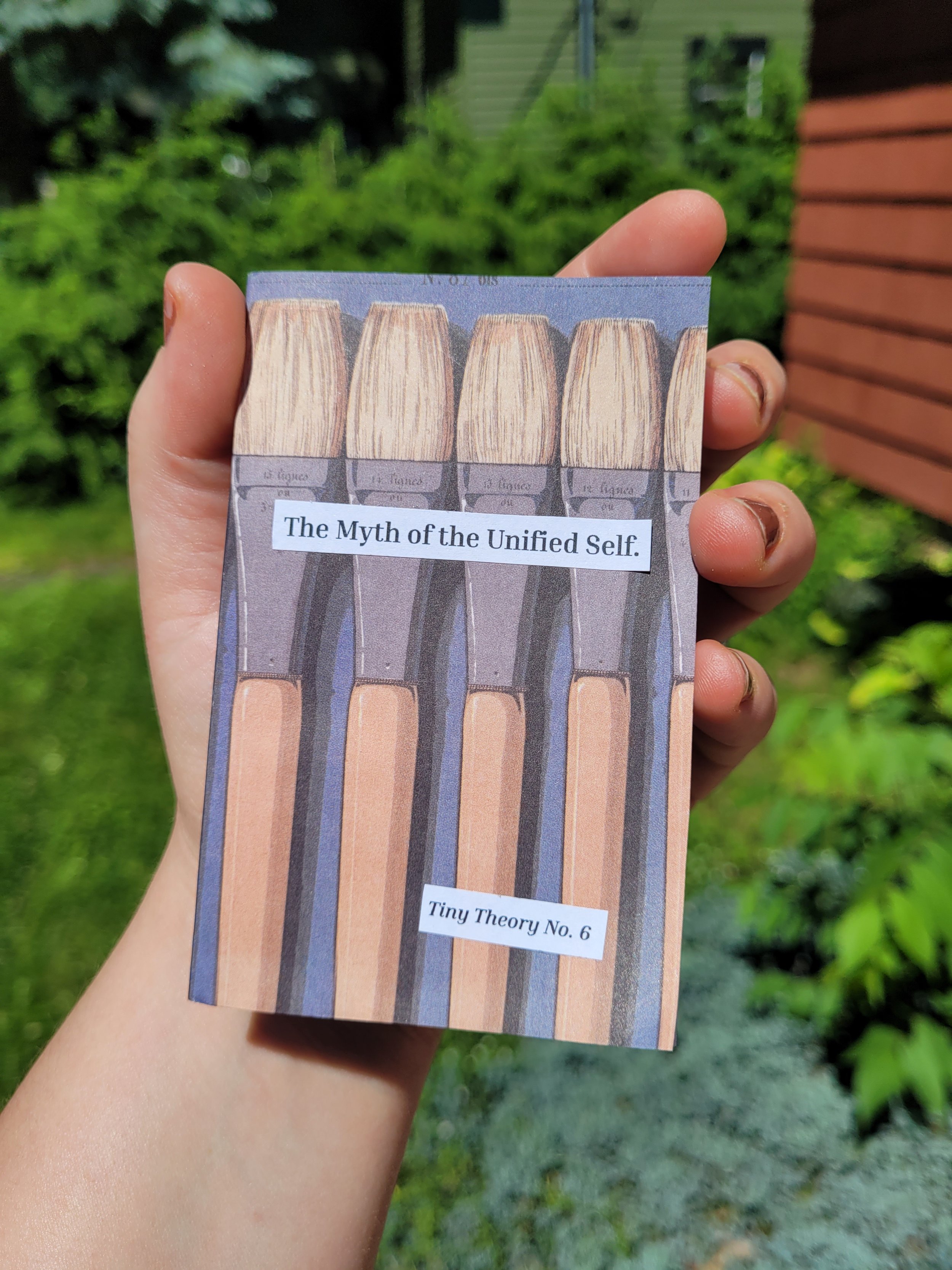 A hand holding a small card with illustrations of different sized paint brushes and the text 'The Myth of the Unified Self.' Tiny Theory No. 6. There is greenery and a building with horizontal siding in the background.