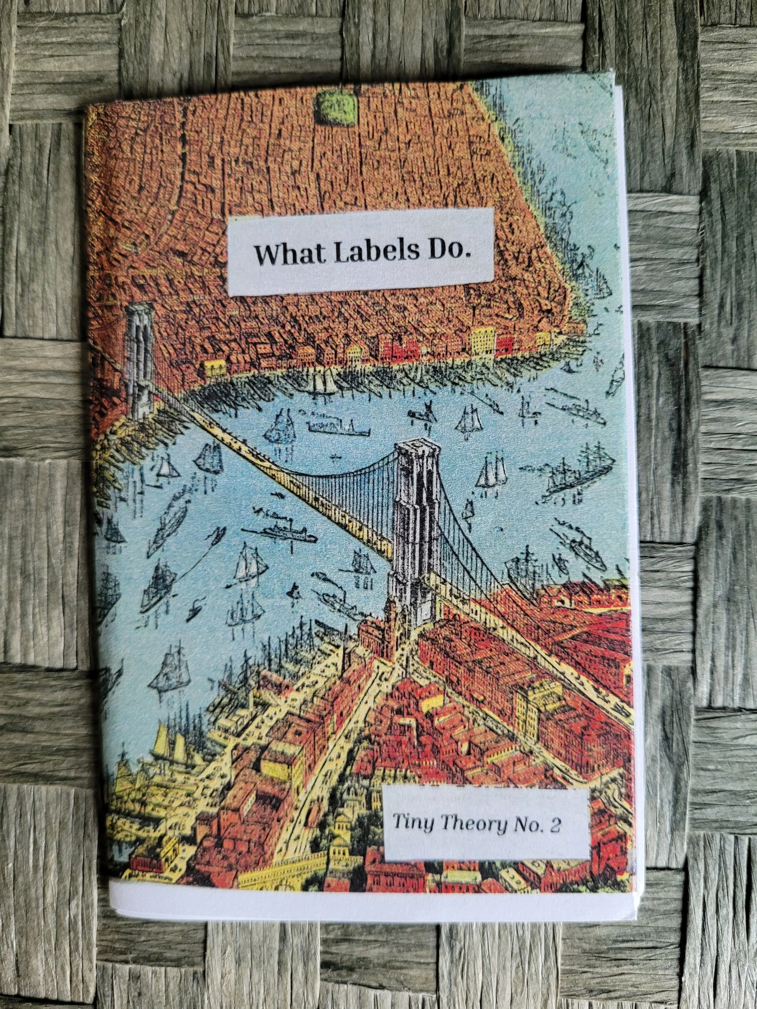 Zine titled 'What Labels Do' with artwork of a red-orange cityscape and a bridge over water, labeled 'Tiny Theory No. 2', placed on a wooden surface.