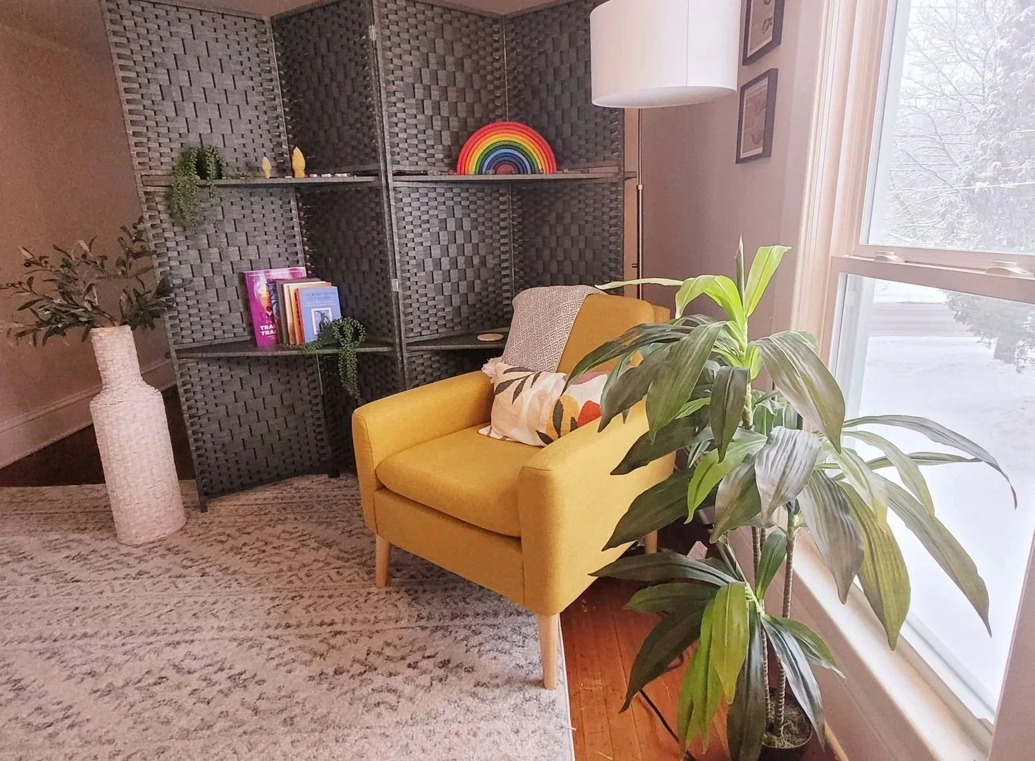 A cozy office with a bright yellow armchair, a large green potted plant next to a window, a woven bookshelf with colorful decorations and books, and a textured beige area rug.