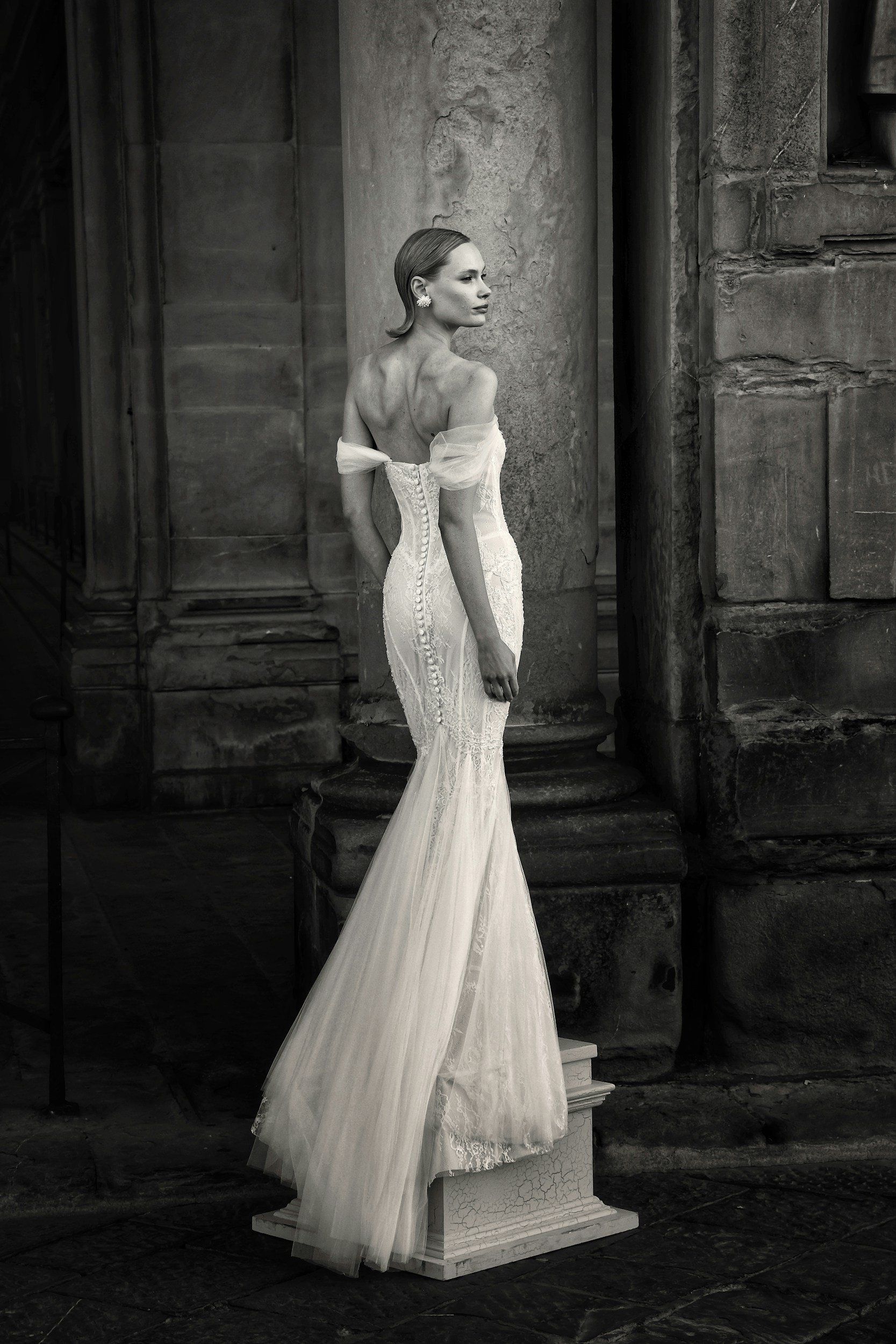 A black-and-white photograph of a woman in a vintage wedding dress leaning against a stone column in an old architectural setting.