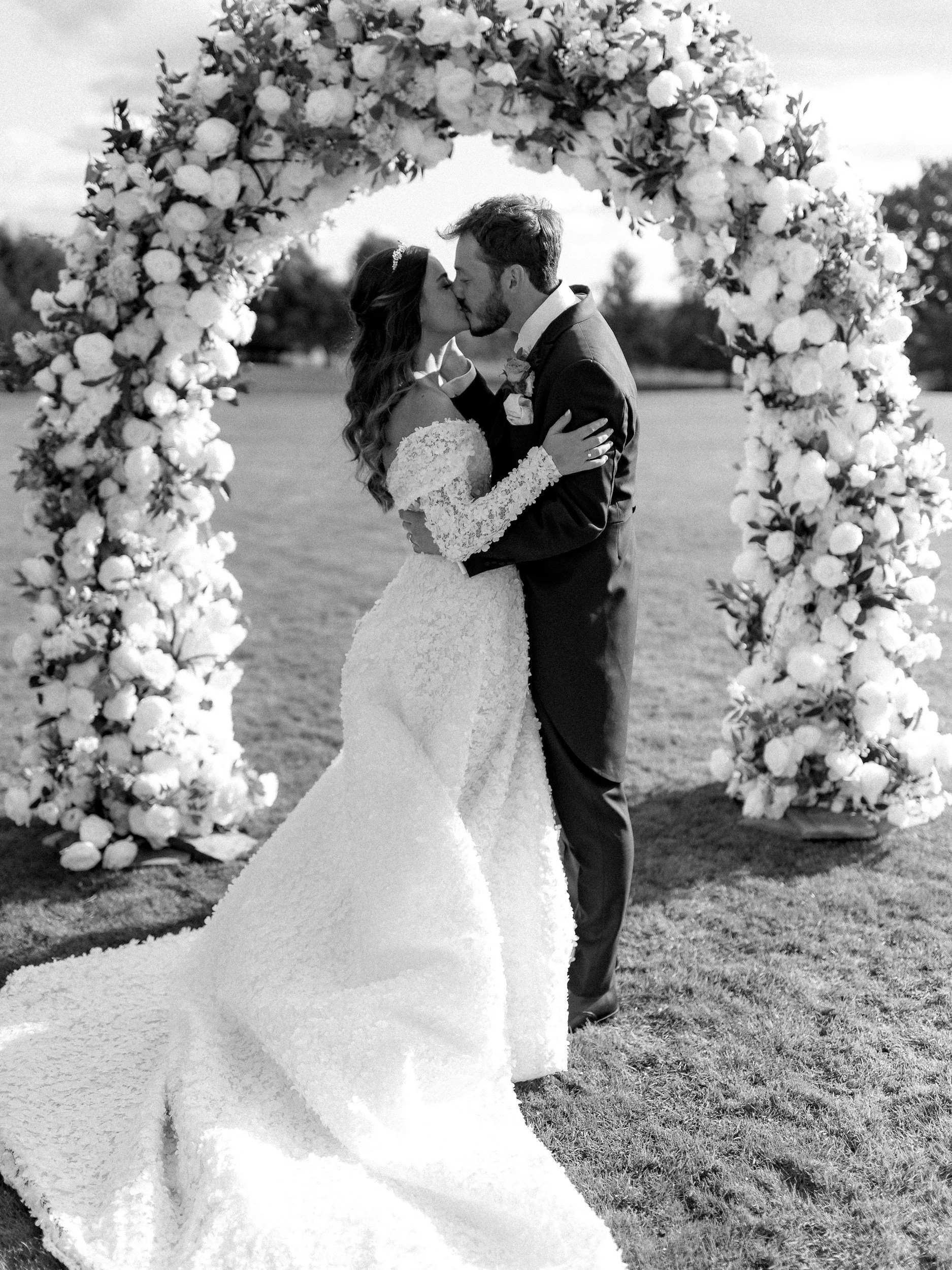 Black and white photo of a bride and groom kissing under a large floral arch on a grassy field in an outdoor wedding setting.