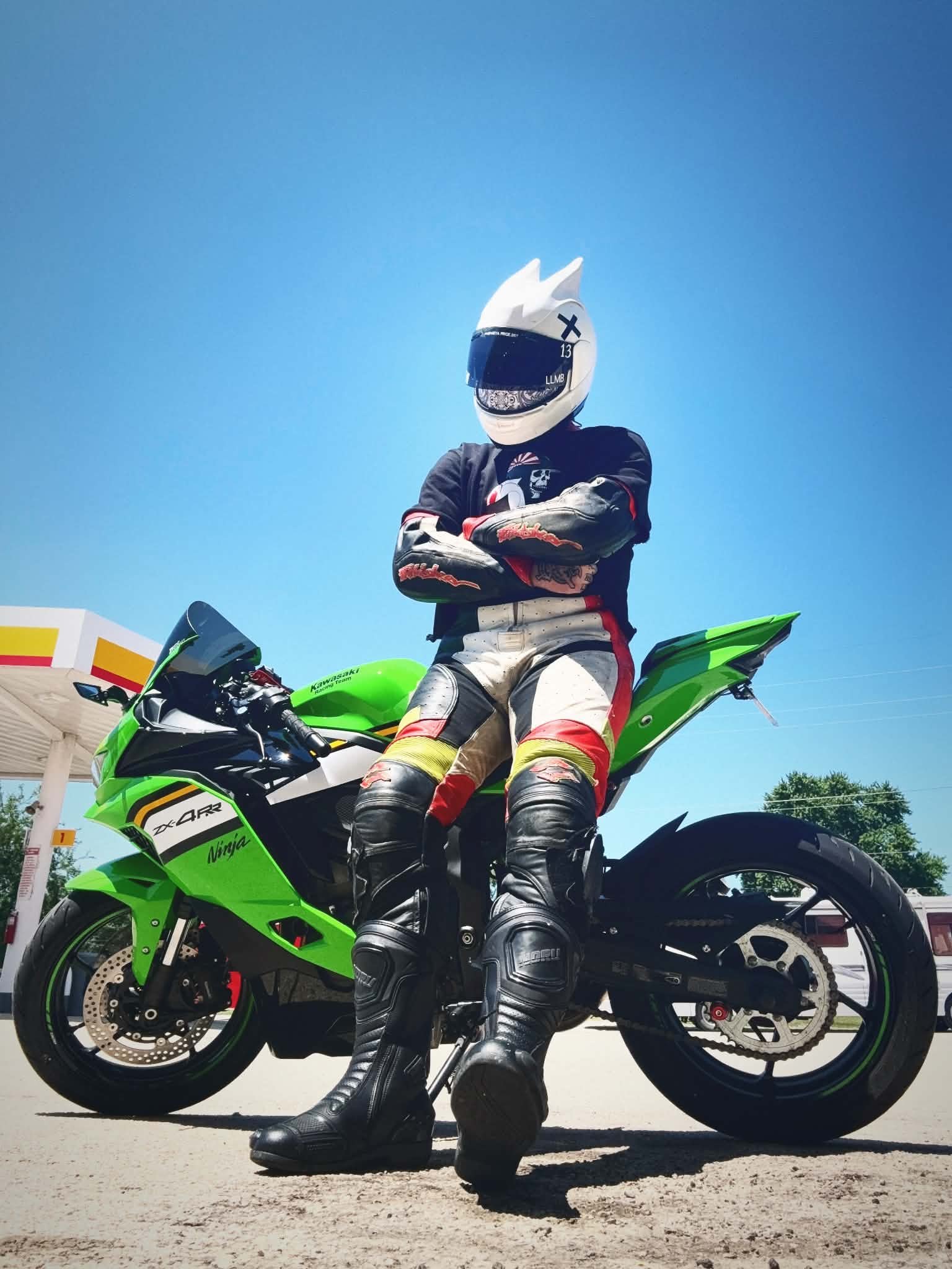 A person in motorcycle racing gear standing next to a green Kawasaki Ninja motorcycle at a gas station on a sunny day.