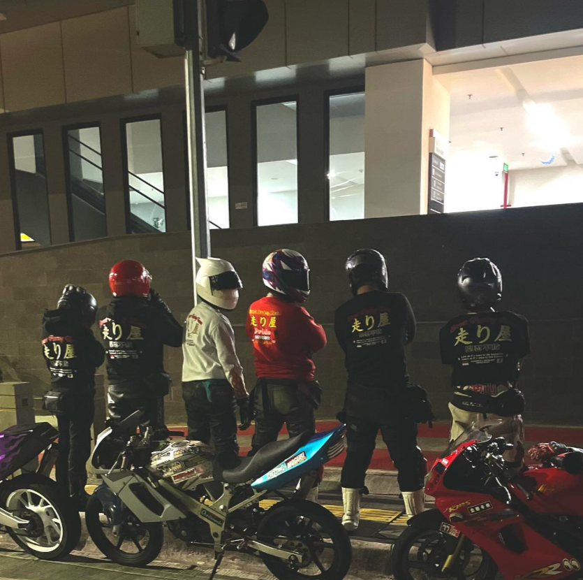 Group of five people wearing racing helmets and black shirts with Japanese characters standing near motorcycles at night.