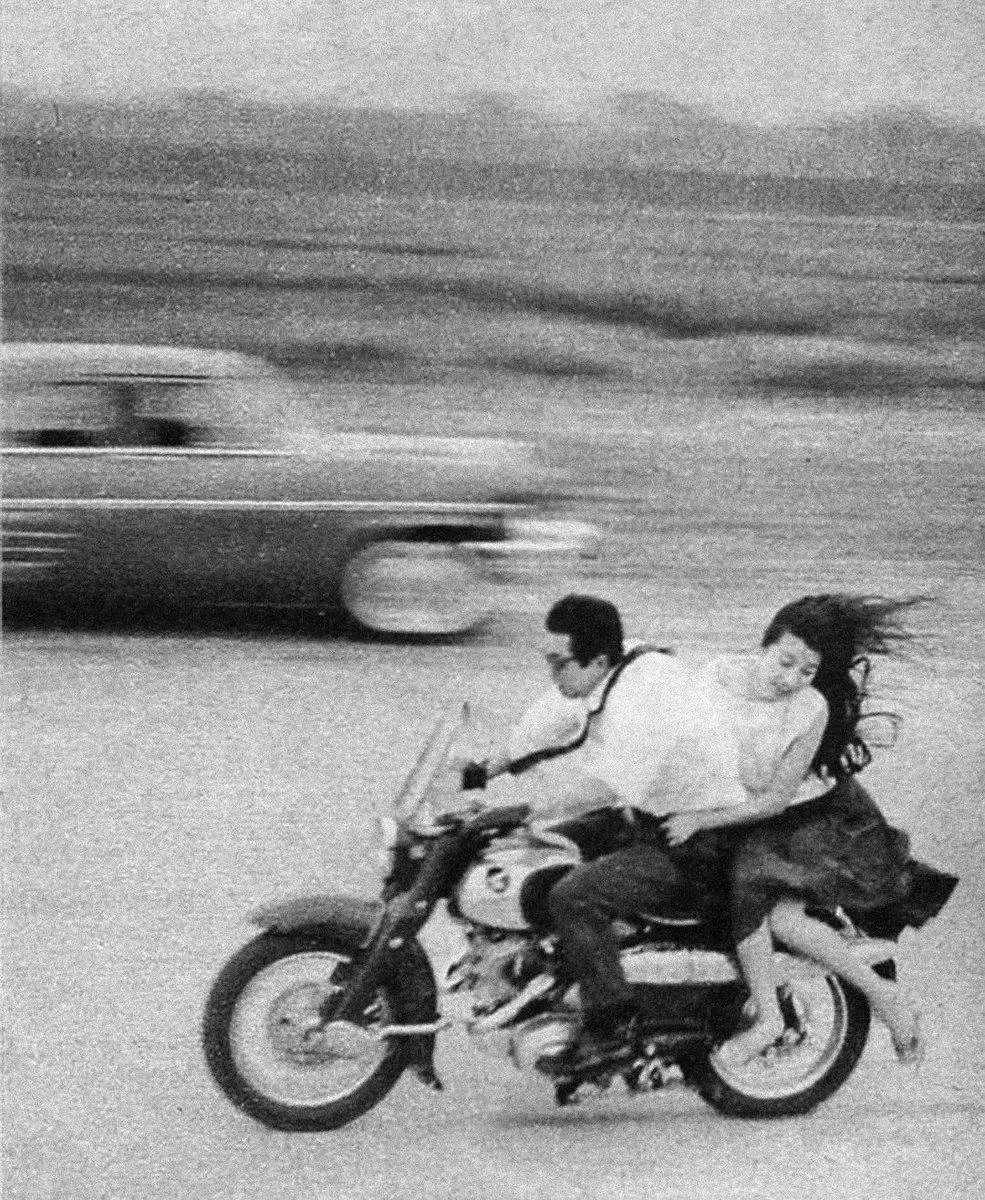 A black and white photo of a man and woman riding a motorcycle at high speed. The woman has long hair flowing behind her and appears to be enjoying the ride, while the man is focused on the road. A car is visible blurred in the background, indicating motion.