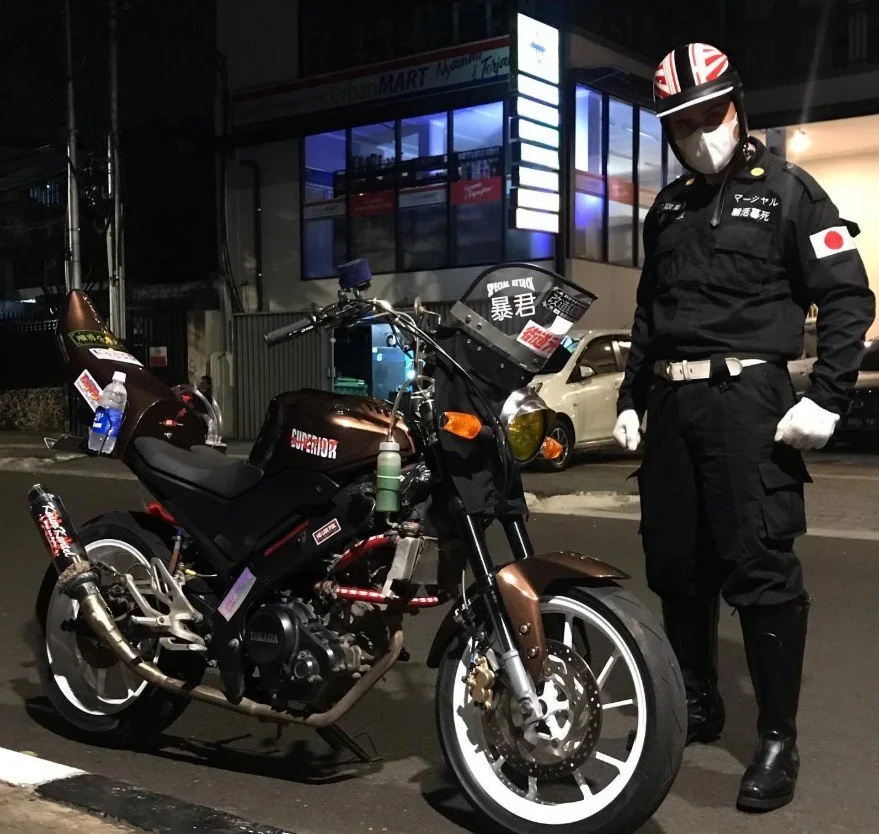 A person dressed in black with Japanese patches, wearing a helmet, face mask, gloves, and boots, stands next to a black motorcycle on a city street at night. The motorcycle has various stickers and accessories.