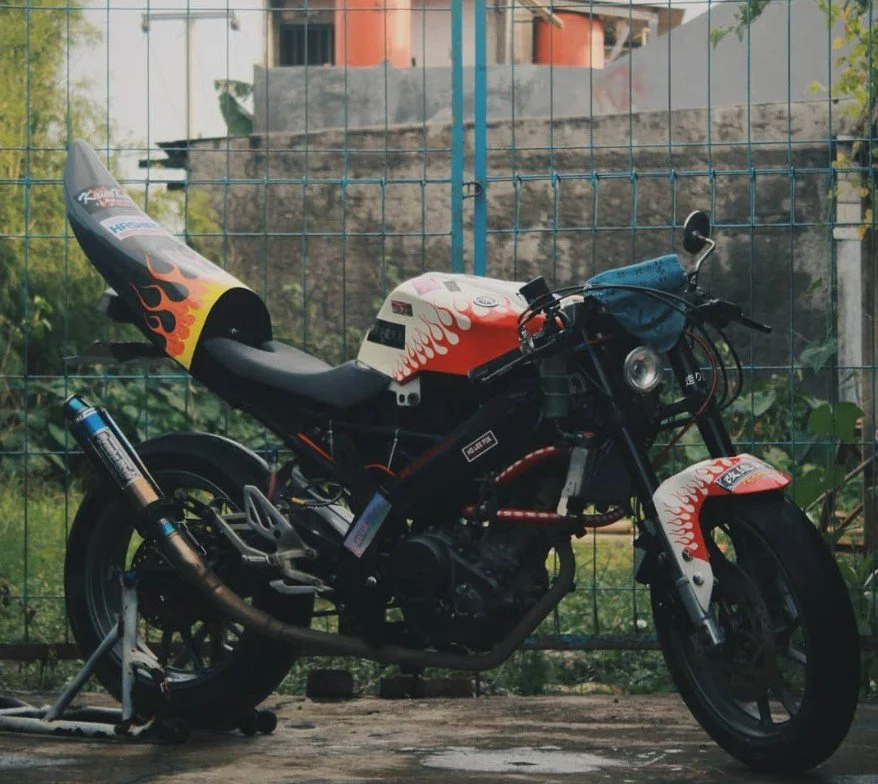A motorcycle with a black frame, flames painted on the white fuel tank, front fender, and rear section, parked in front of a metal fence with plants and a building in the background.