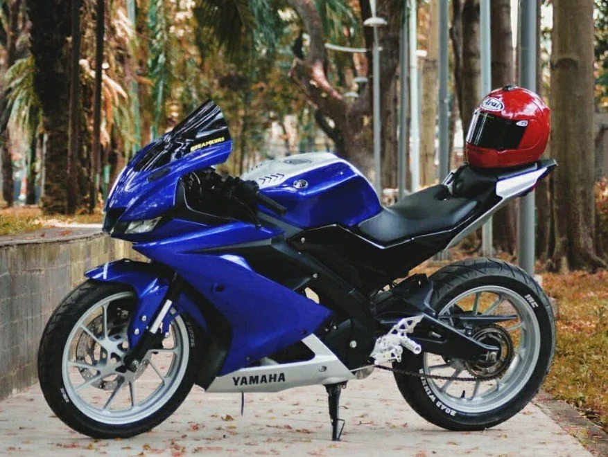 Blue Yamaha sport motorcycle with a red helmet on the seat, parked on a concrete path in a wooded area.