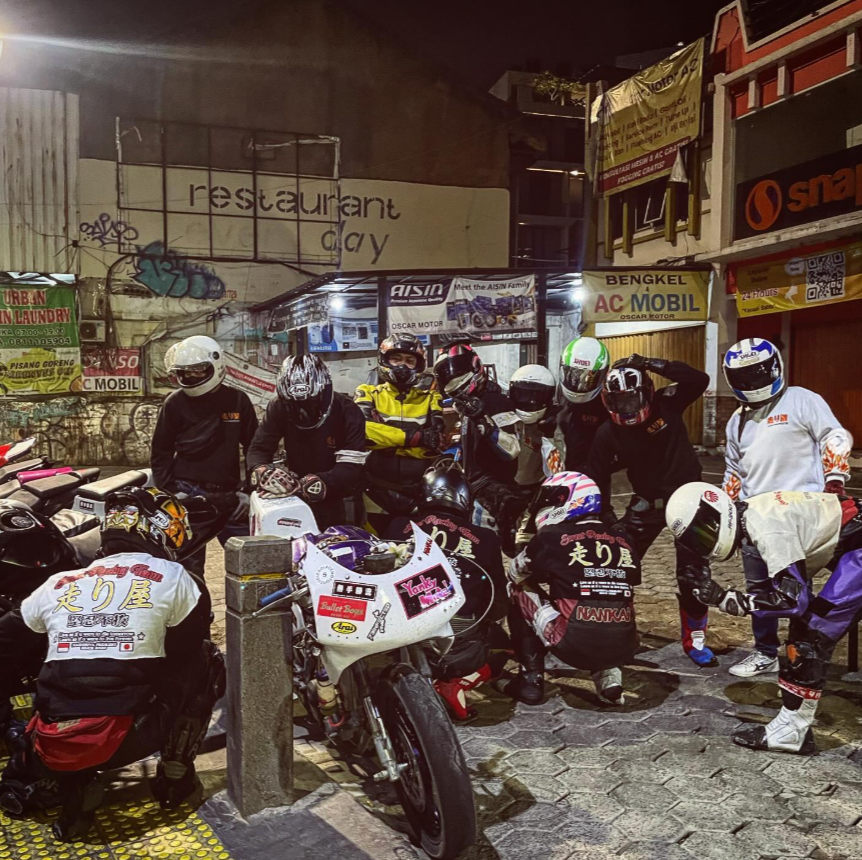 Group of motorcyclists in racing gear gathered around motorcycles on a city street at night.